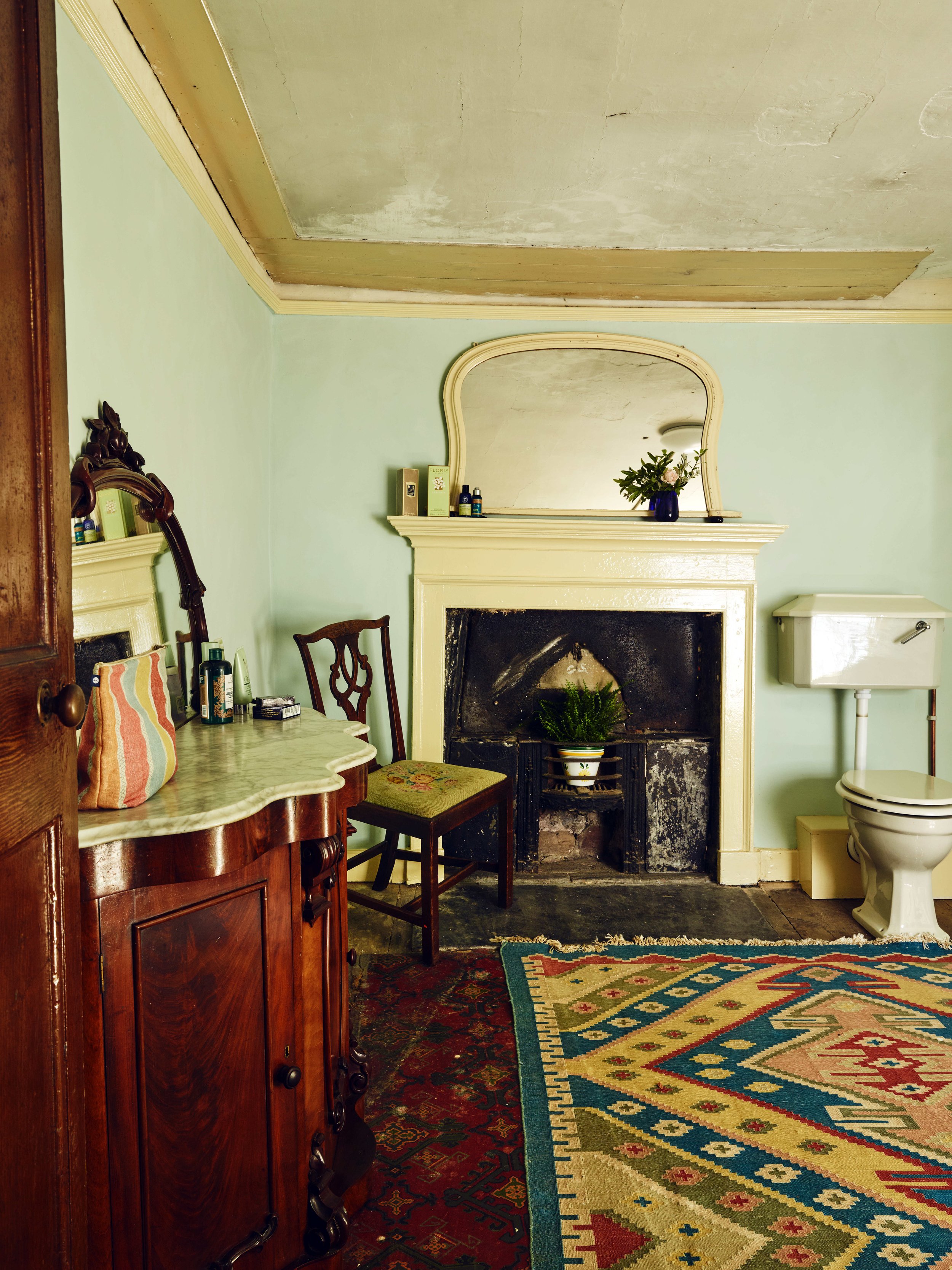 A living room in a 16th century house in Cornwall with a mantel, mirror, and fireplace, decorated with flowers and bottles. There is a wooden cabinet, a floral chair, and a colorful patterned rug. A small section of a toilet is visible in the corner.