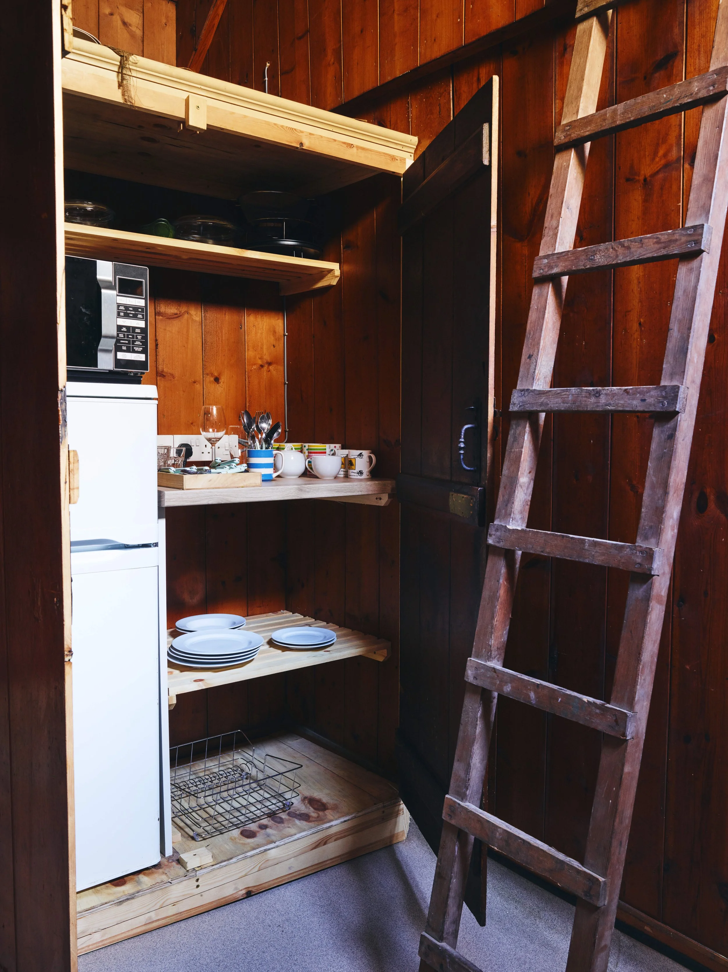 Wooden kitchen storage with shelves holding dishes, cups, utensils, a microwave, and a small fridge, alongside a wooden ladder leaned against a wood-paneled wall.