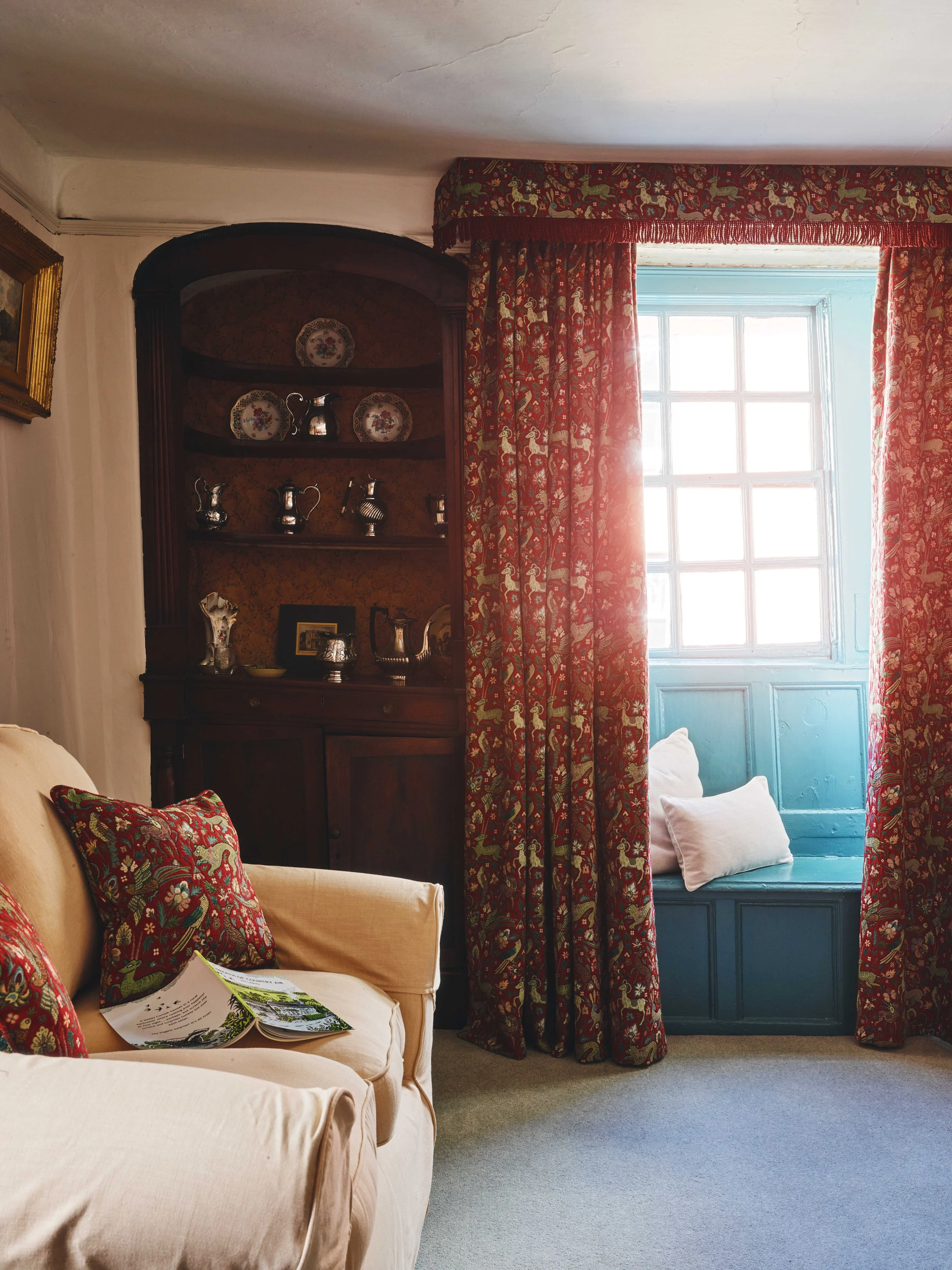 A living room corner with a cream sofa adorned with red and green patterned pillows. A wooden cabinet displays vintage silver tea sets and decorative plates. A large window with blue-framed panes is covered by red patterned curtains, with a window se