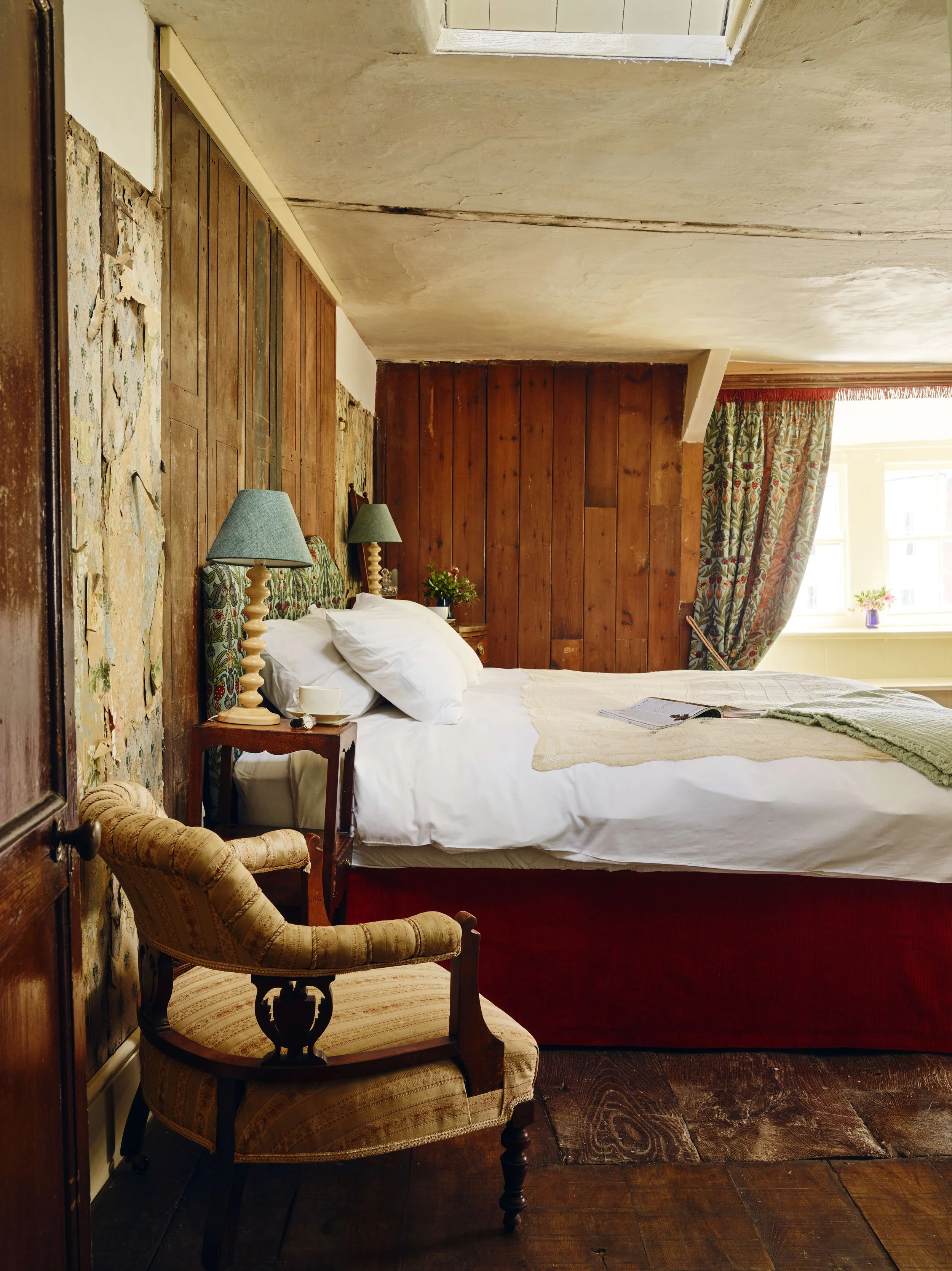 A bedroom with a large bed, wooden paneled walls, and a window with floral curtains. The bed has white linens and a green blanket, with a book and glasses on top. There are two blue lamps on bedside tables and a vintage armchair beside the bed.