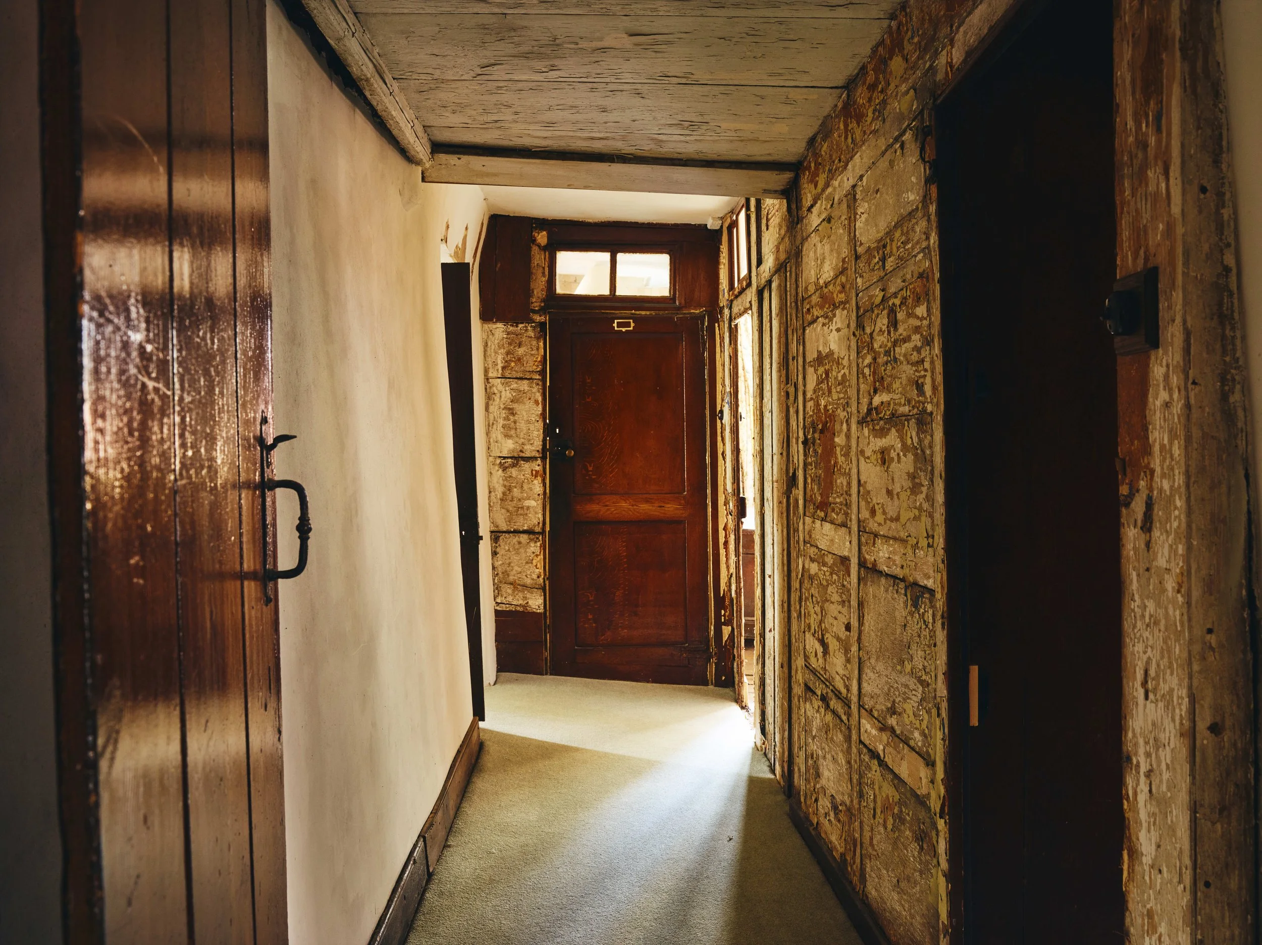 A hallway in a 16th century house in Cornwall with worn, peeling wall paint and aged wooden doors at the end, illuminated by natural light.