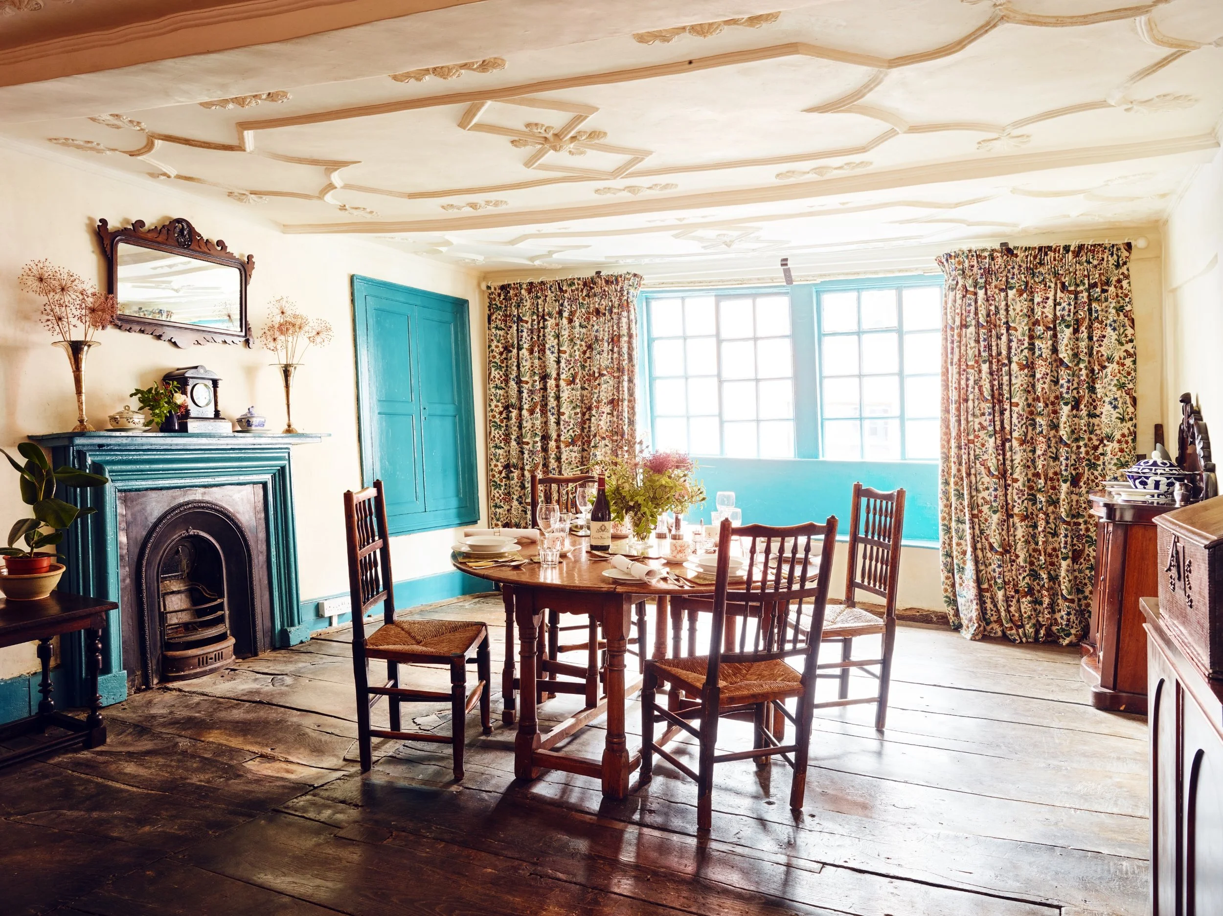 A historic dining room with a round wooden table set with dishes, glasses, and a bottle of wine, surrounded by four wooden chairs, with floral curtains and a blue window in the background, and an ornate fireplace on the left side.