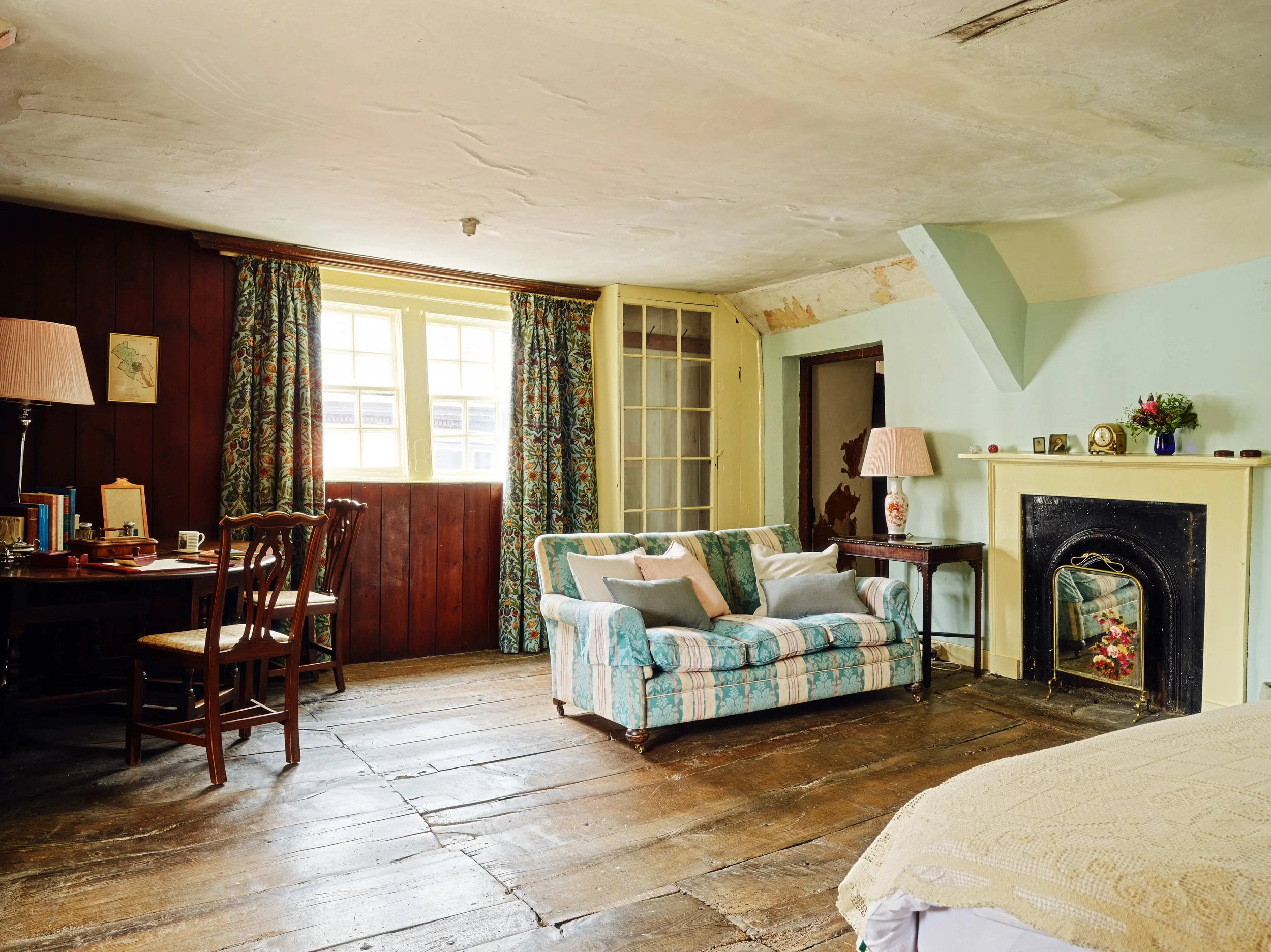 A living room in a 16th century house in Launceston with wooden floors, a patterned sofa with multiple pillows, a fireplace with a mirror, a table with a lamp, curtains, and a window letting in natural light.