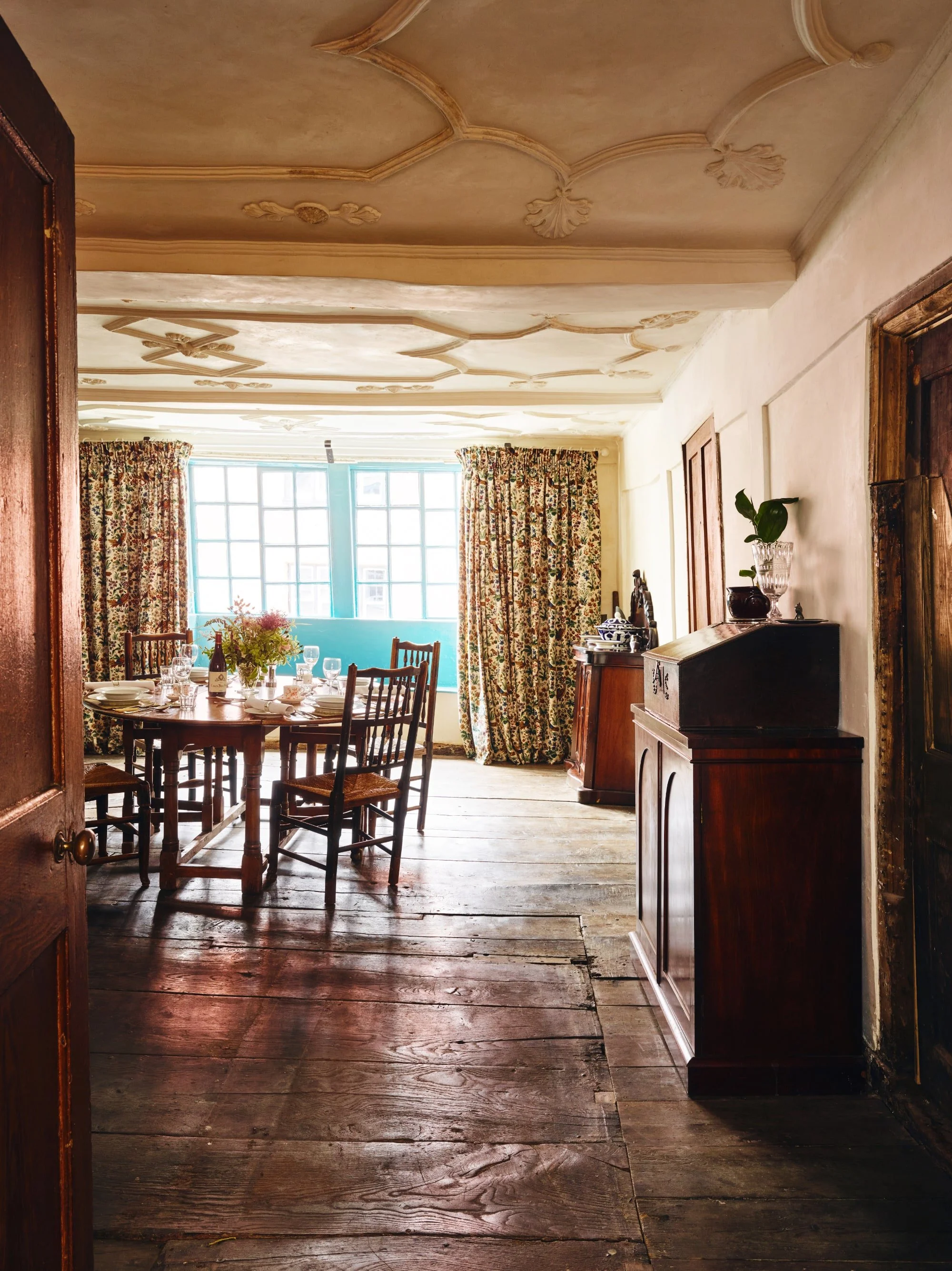 A dining room with a wooden dining table set with plates, glasses, and a wine bottle, surrounded by wooden chairs, with floral curtains and a large window letting in natural light, hardwood floors, and vintage furniture.