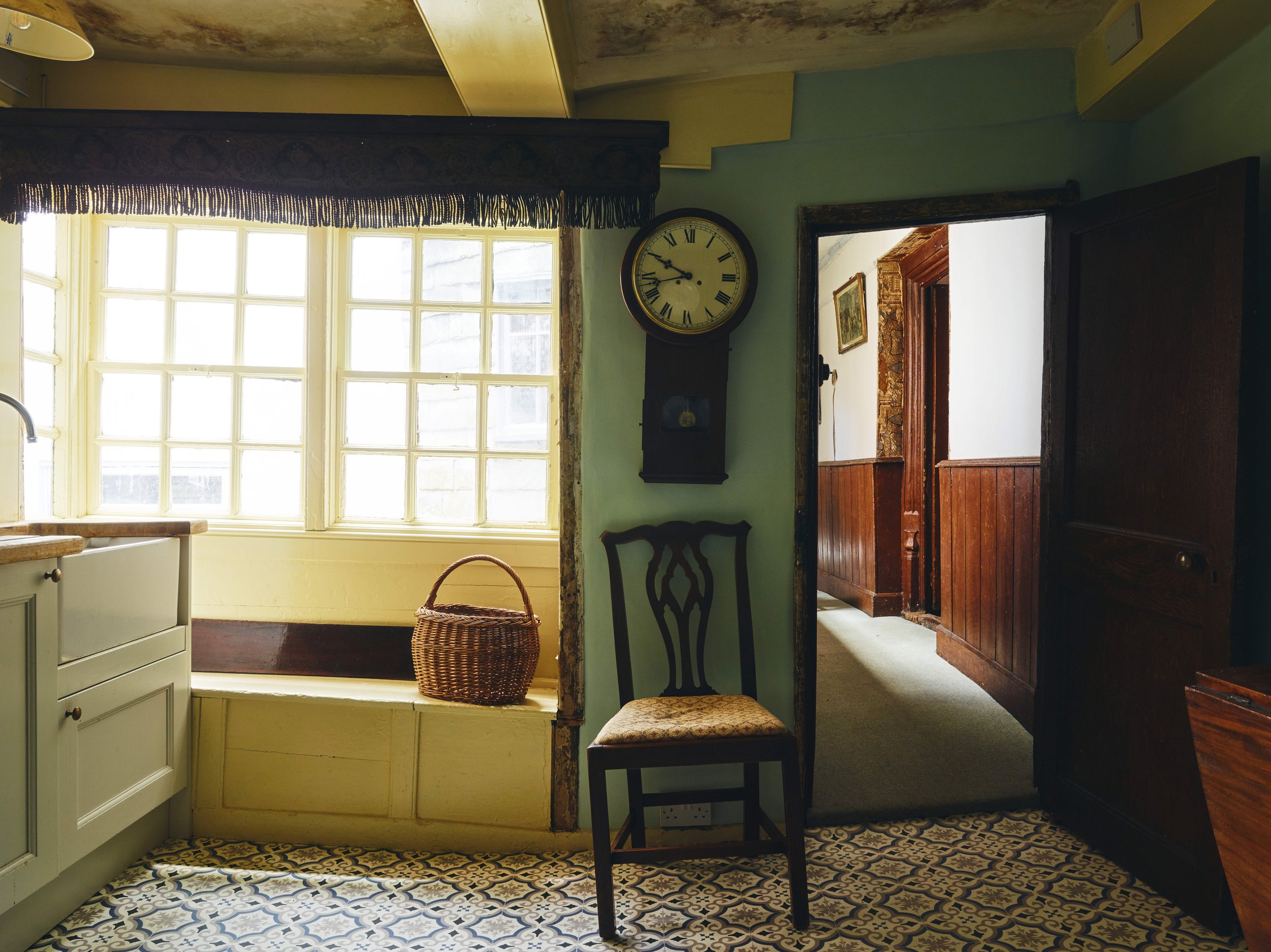 16th century house in Launceston - kitchen with patterned tile floor, a large window with a bench and basket, a wall clock, a wooden chair, and a doorway leading to a corridor with wood-paneled walls.
