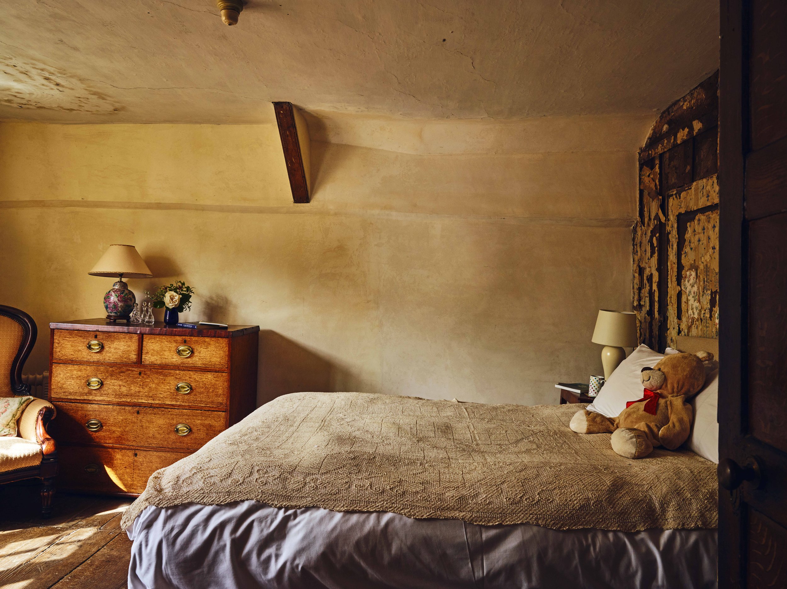 A bedroom with a large bed featuring a teddy bear on top, a bedside table with a lamp and mug, a dresser with a flower arrangement and decorative glassware, an armchair with floral upholstery, and an ornate wall decoration above the bed. The room has