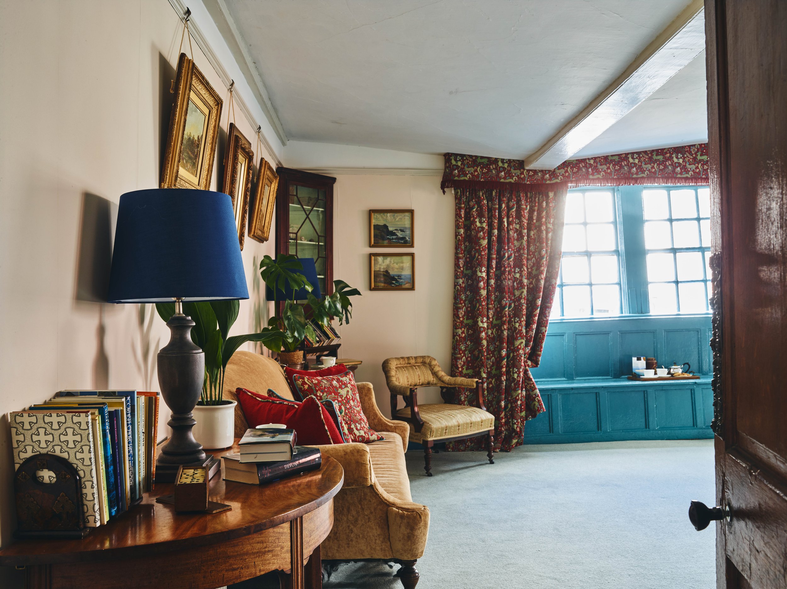 A historic living room with antique furniture, a beige sofa with red cushions, a wooden table with books and a lamp, a patterned armchair, a wall with framed landscape paintings, and a window with floral curtains and teal woodwork.