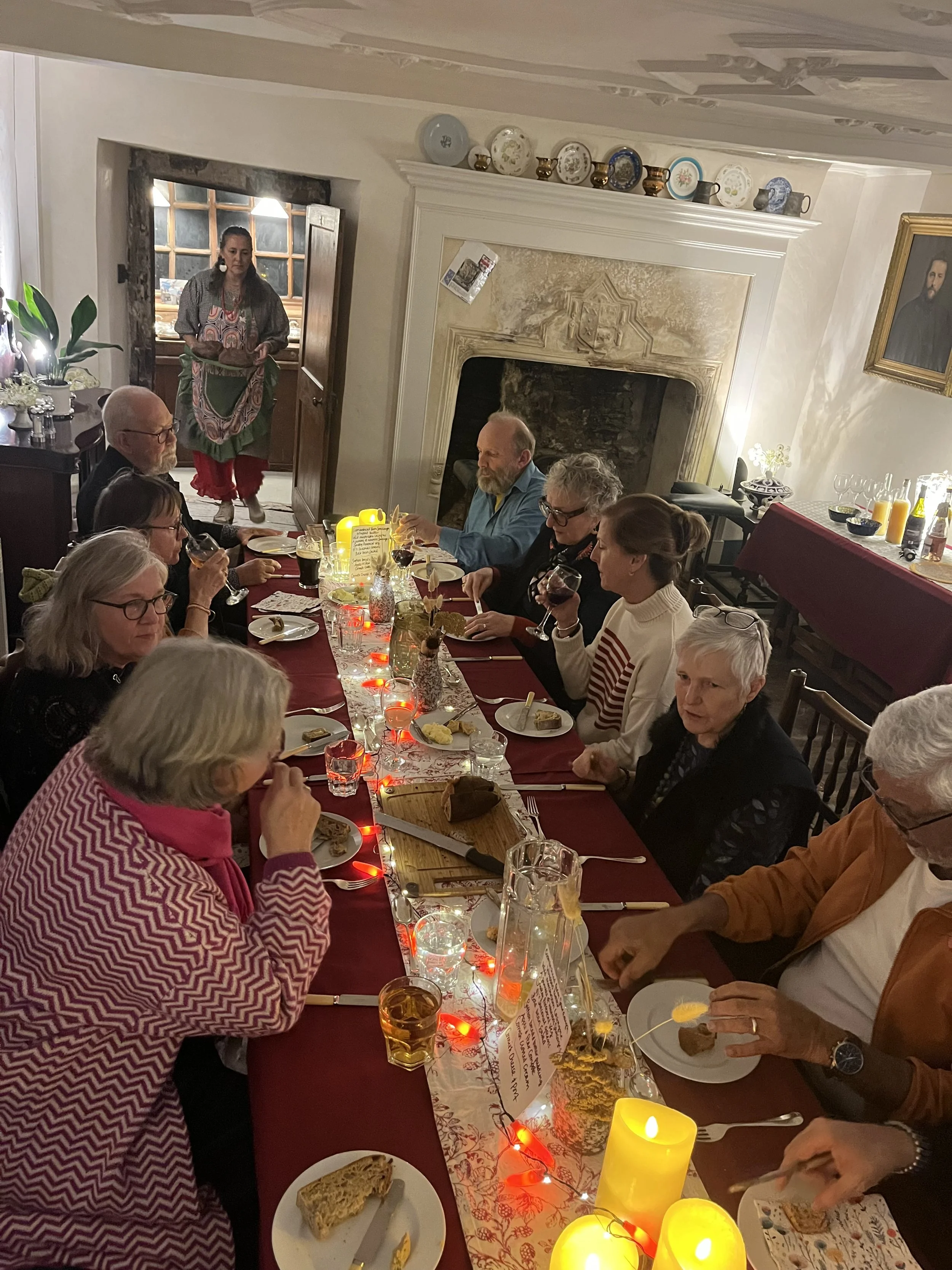 People gathered for a dining club at a long table decorated with candles and a table runner in a historic, illuminated room with a fireplace and a portrait on the wall.
