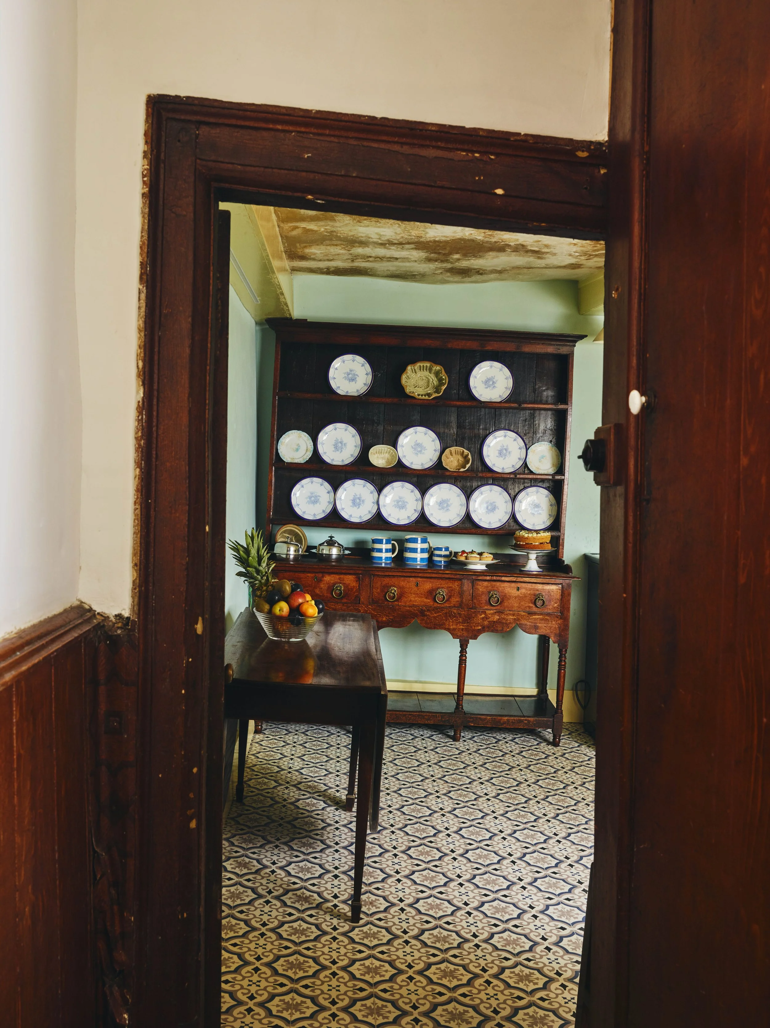 A view through a wooden door frame into a vintage kitchen with a dark wooden sideboard, a corner shelf with blue and white plates, a bowl of fruit, and a tiled floor.
