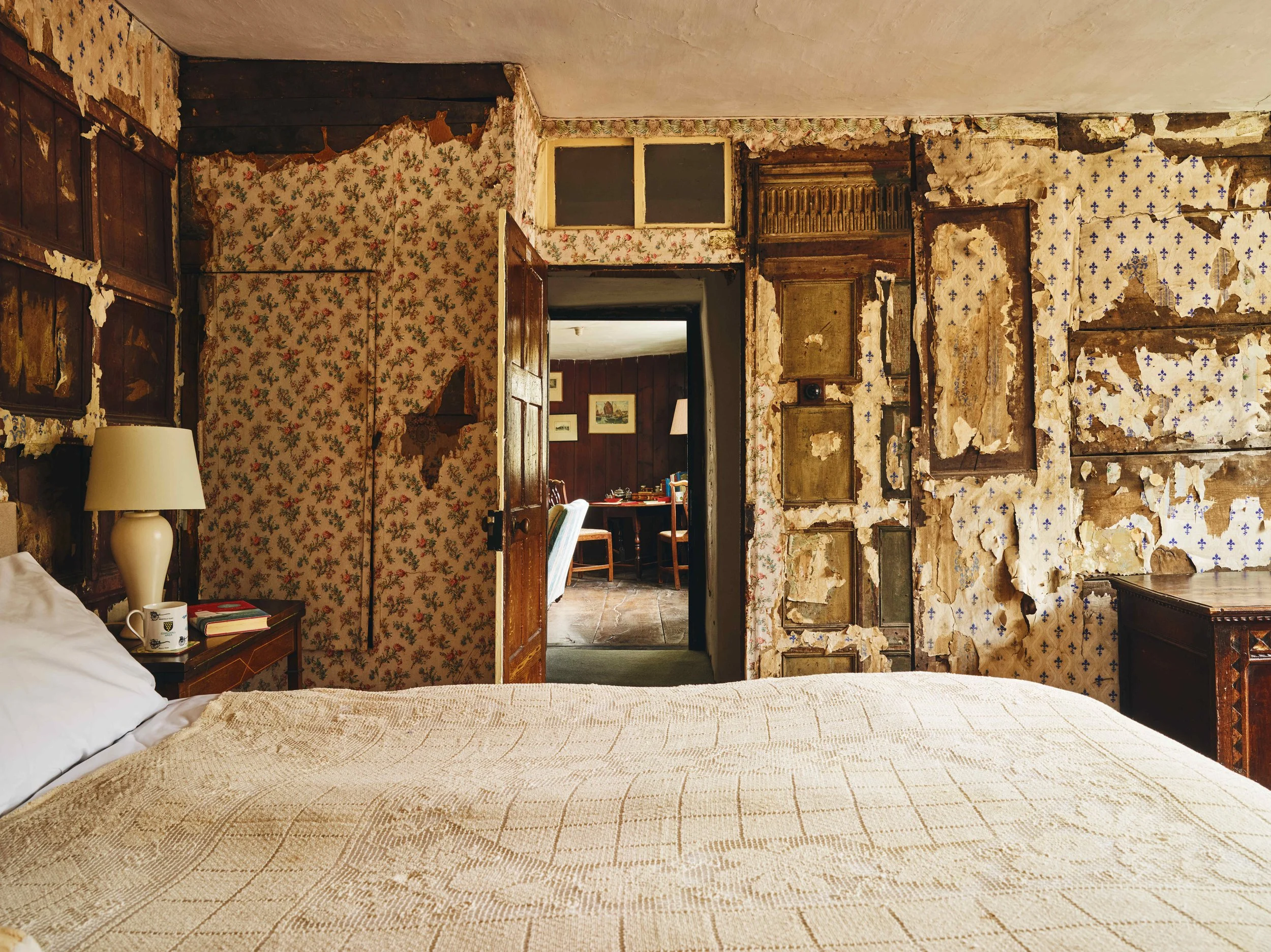 A bedroom with a vintage aesthetic featuring a bed with a cream-colored quilt, a nightstand with a white lamp, a mug, and books; worn floral wallpaper and exposed patches of wallpaper on the walls; a doorway leading to a dining room with wooden panel