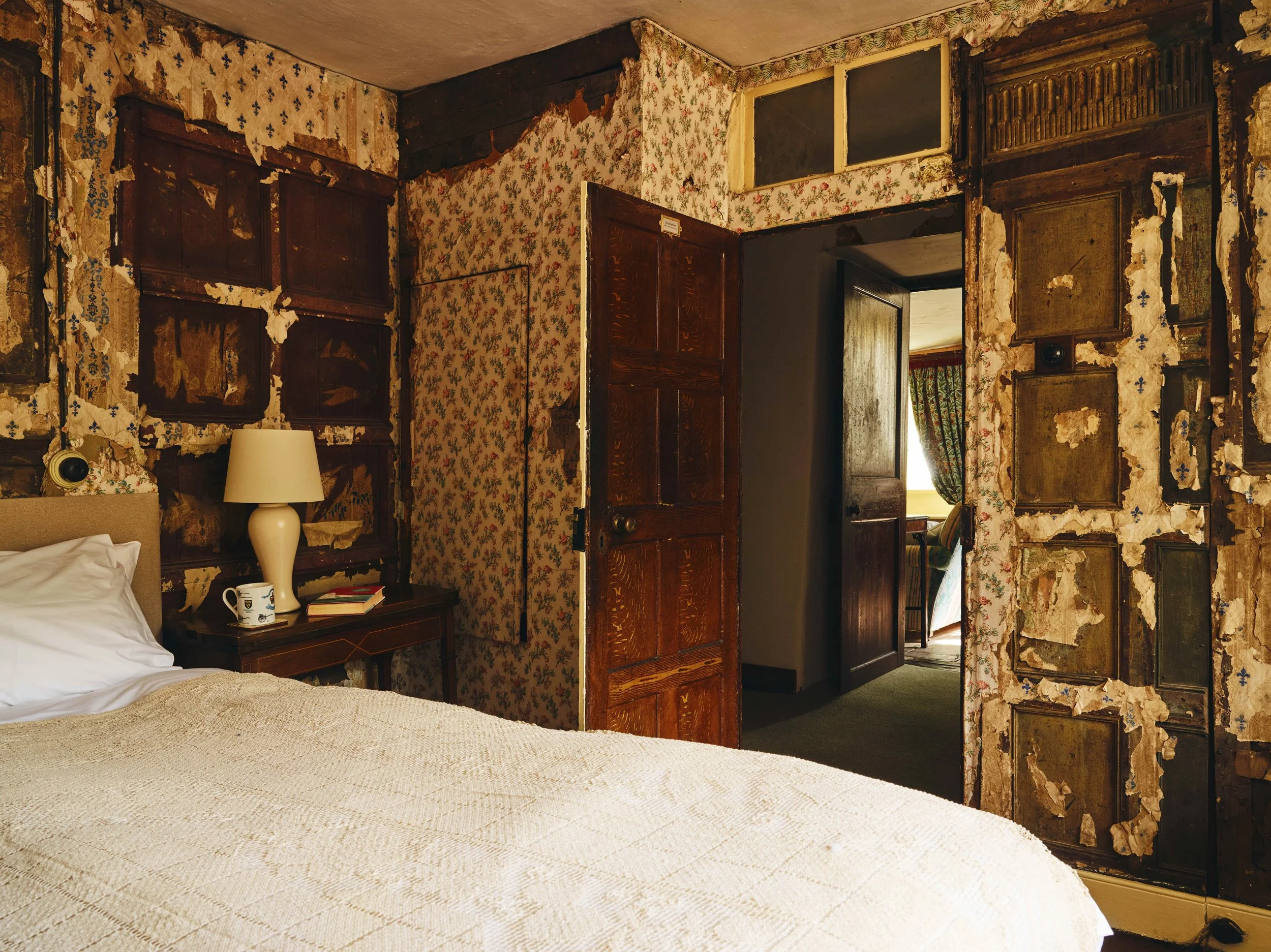 A bedroom 16th century house in Cornwall with peeling floral wallpaper, a bed with white linens, a bedside table with a lamp, books, and a mug, and an open wooden door leading to another room with green curtains and a window.