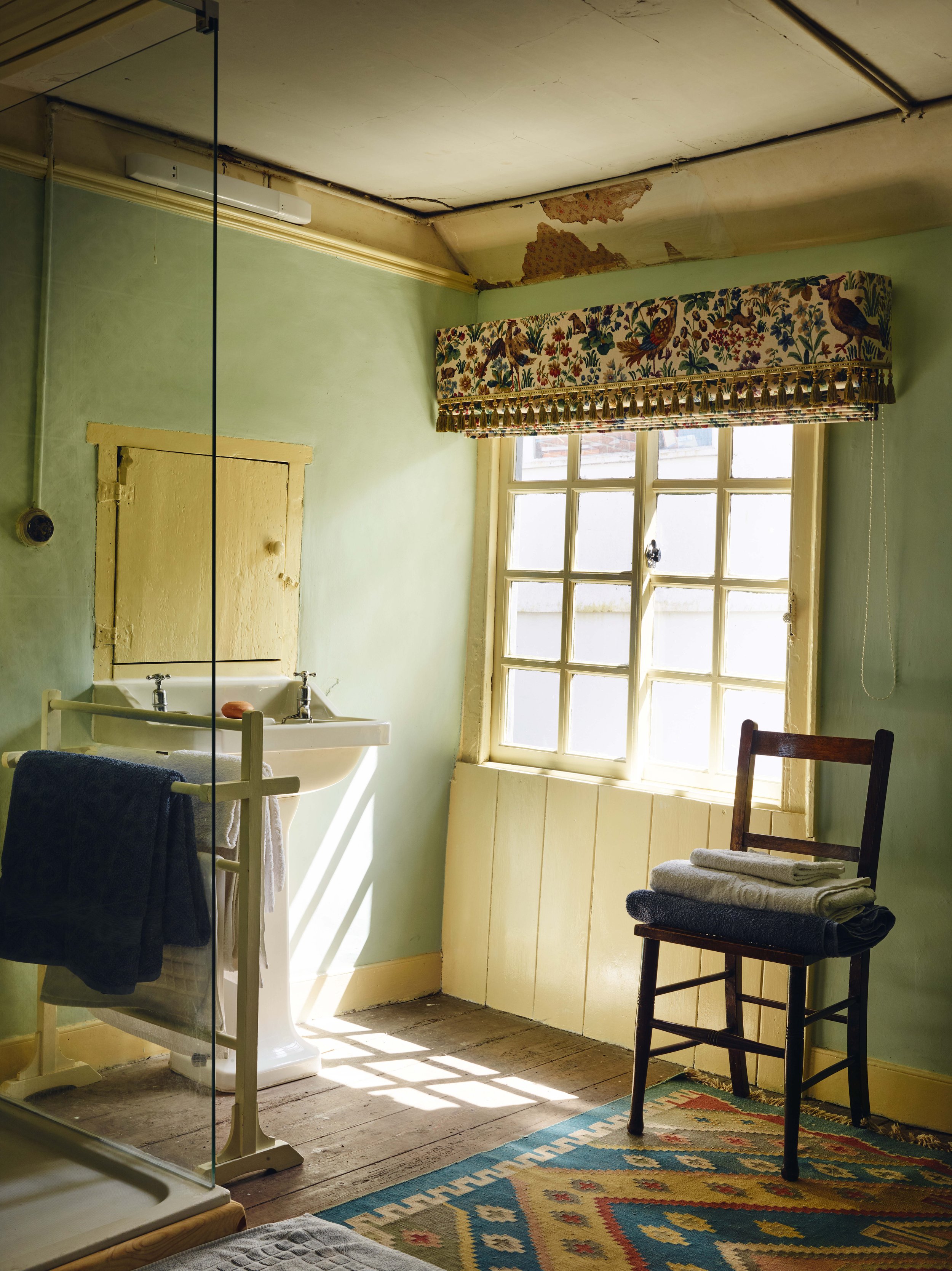 A bathroom with a yellow framed sink, a window with floral valance, a wooden chair with folded towels, an area rug, and peeling paint on the ceiling.