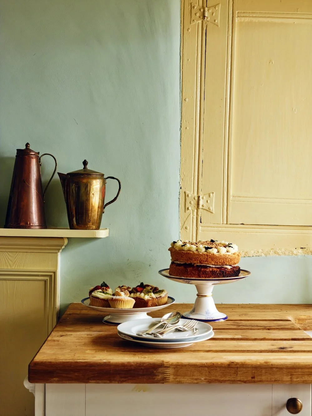 A wooden table with desserts including a layered cake on a cake stand, cupcakes with frosting, and a stack of plates with silverware, next to a wall with a partial yellow wooden door and a green wall, with two vintage metal containers on a shelf.