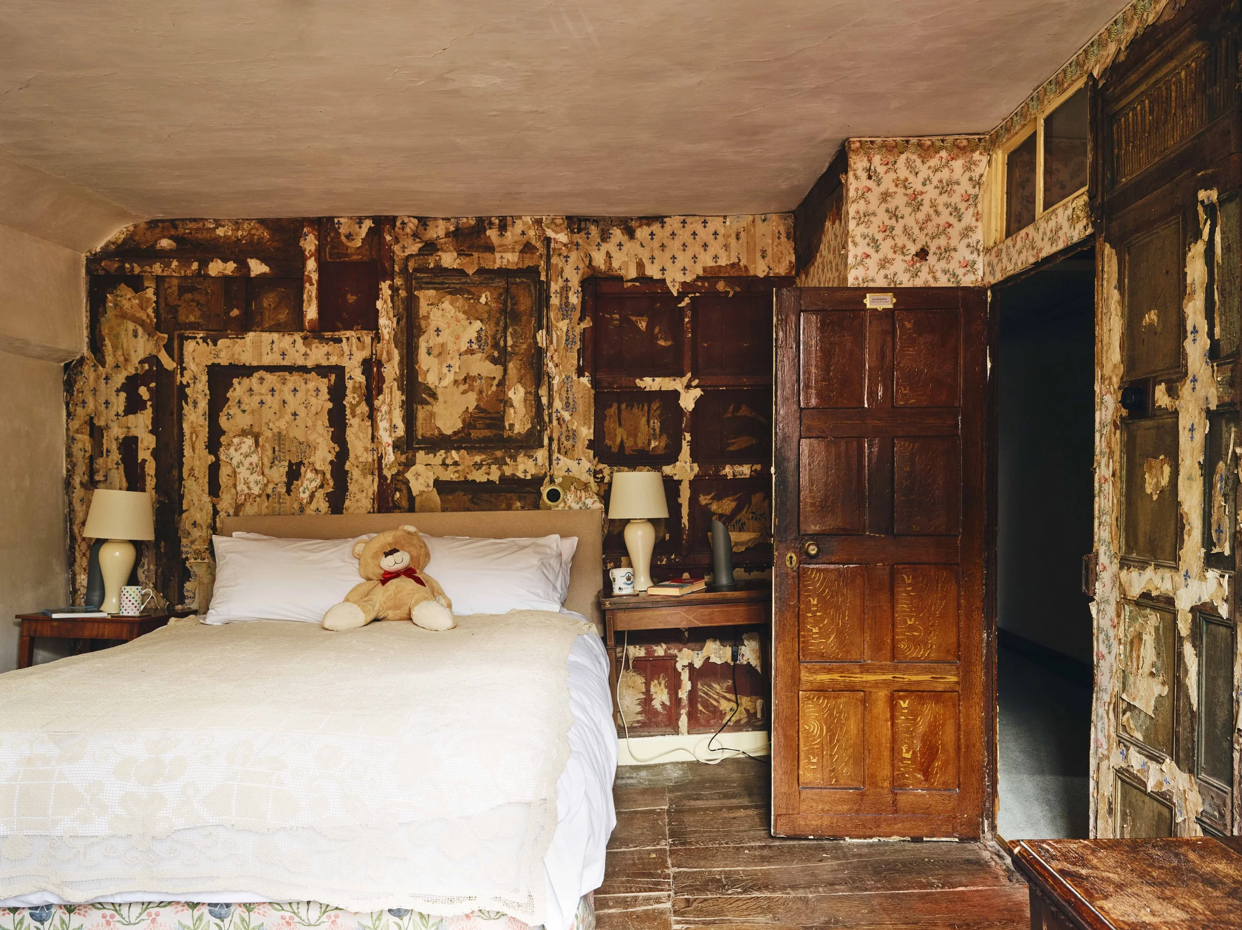 A bedroom 16th century house in Launceston with a bed featuring a teddy bear, two nightstands with lamps, and a wall decorated with peeling wallpaper and wooden panels in a rustic style.