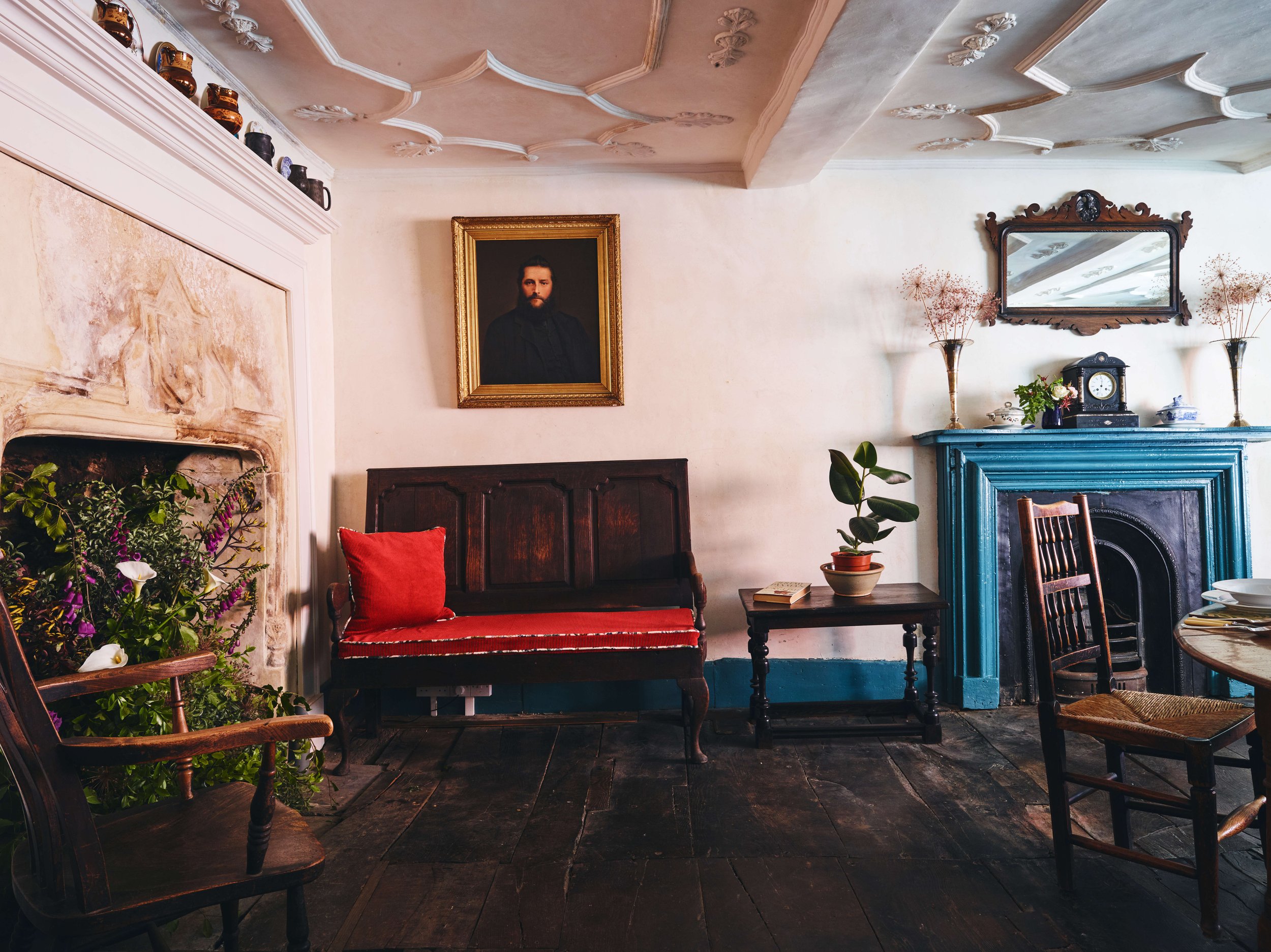 A living room with vintage furniture, including a dark wood bench with red cushion, a wooden side table with a potted plant, and a round dining table with chairs. There is a large portrait of a man with a beard hanging on the wall above the bench. Th