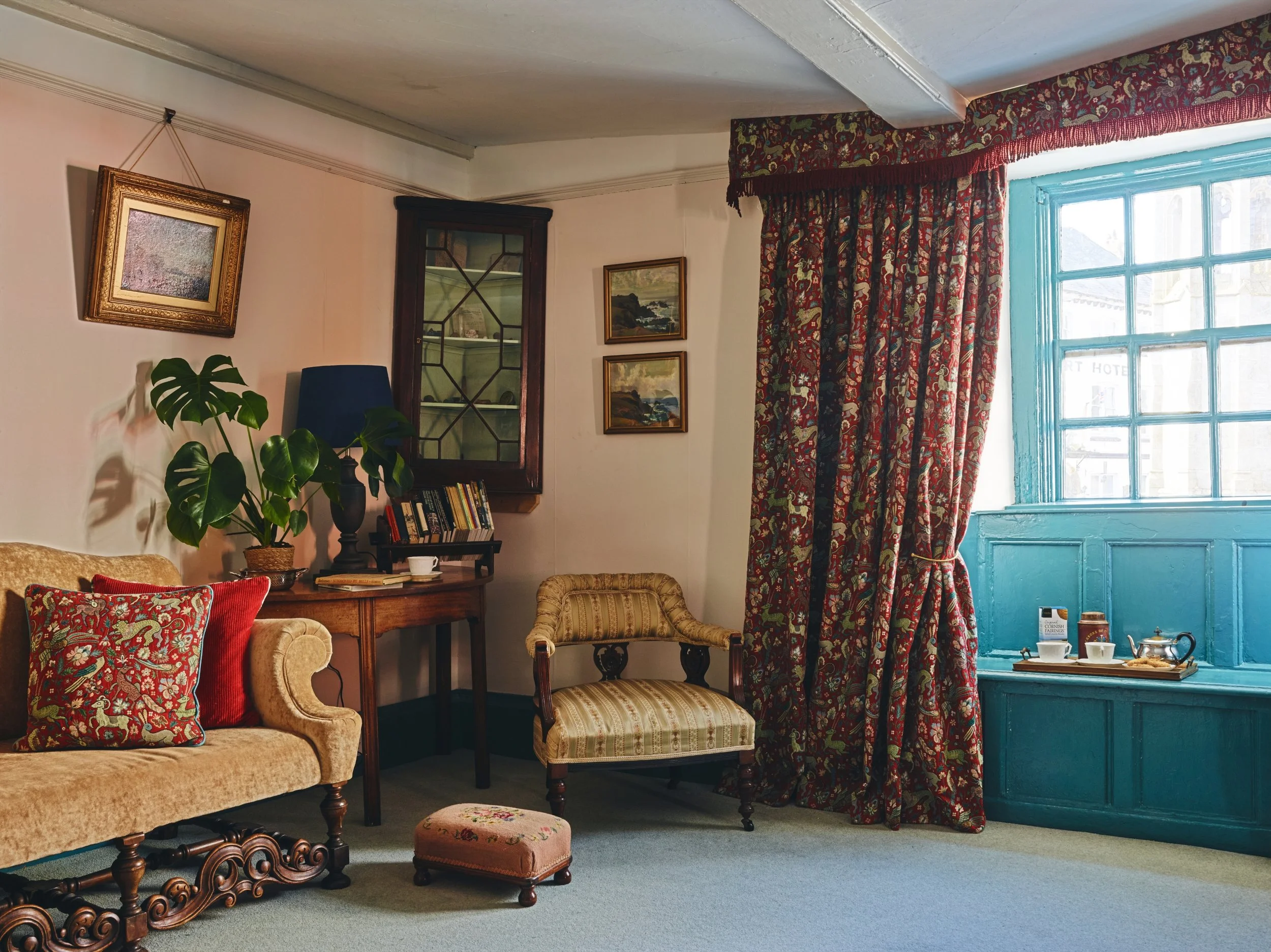 A historic living room with a beige sofa with red pillows, a wooden side table with a plant and books, an upholstered armchair, a window with blue framing and floral curtains, and a small ottoman with floral embroidery.