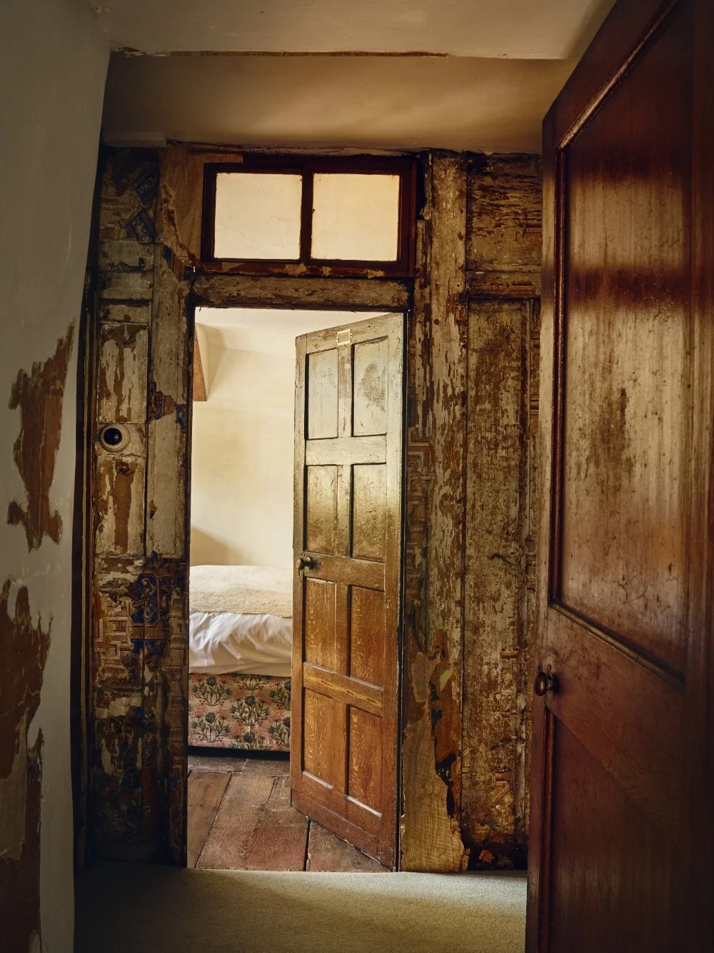 Historic, weathered wooden door slightly open, leading into a room with a bed and a white wall, with peeling paint and exposed wood framing around the door.