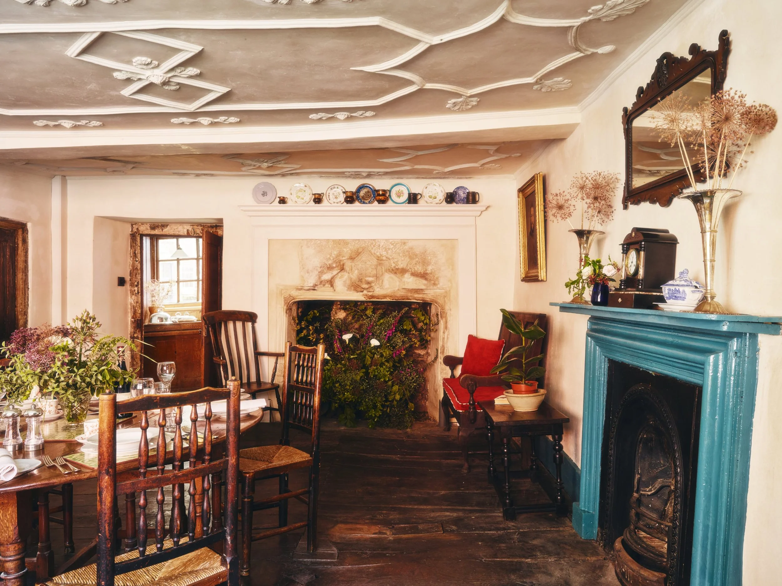 A historic dining room with a wooden table and chairs, floral arrangements, a fireplace with plants, a mirror, and decorative plates on the mantel, with muted cream-coloured walls and a textured ceiling.
