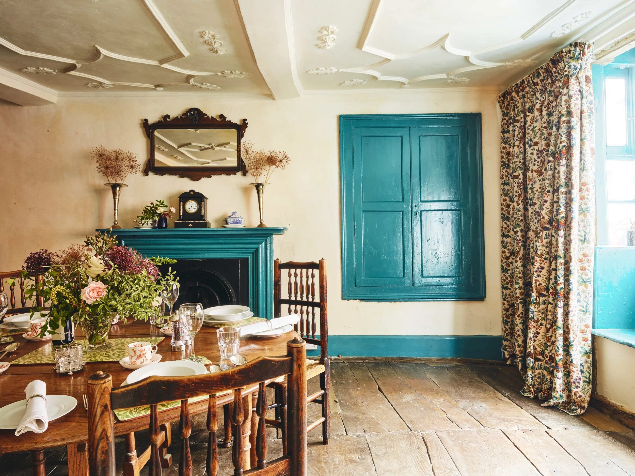 A dining room with a wooden table set for a meal, decorated with a vase of flowers, surrounded by wooden chairs. The room features a blue fireplace mantel, a mantle clock, and decorative vases with dried flowers. There is a mirrored picture frame abo