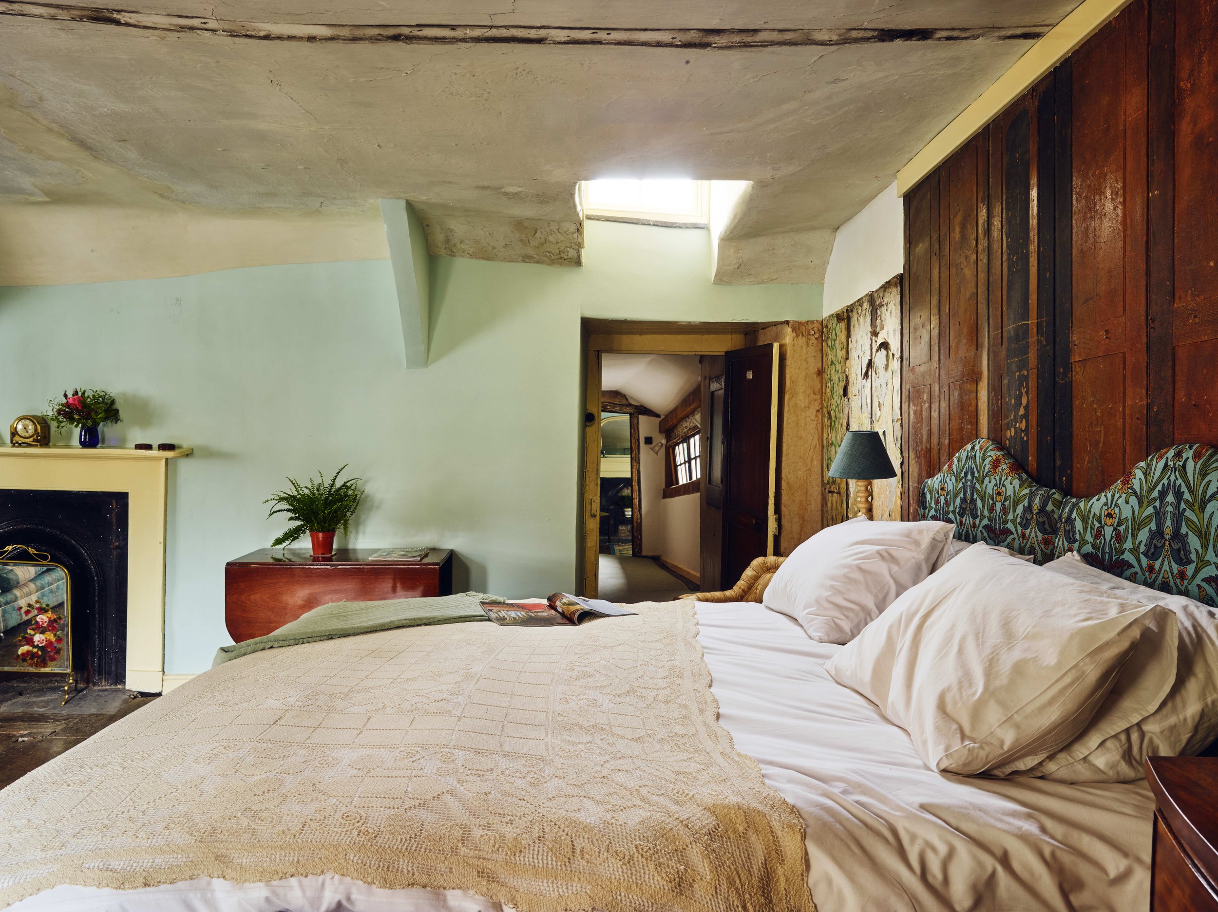 Historic bedroom with a white bed, quilt, and pillows, wooden headboard, bedside lamp, small table with plants, and a fireplace, with a hallway leading to another room.