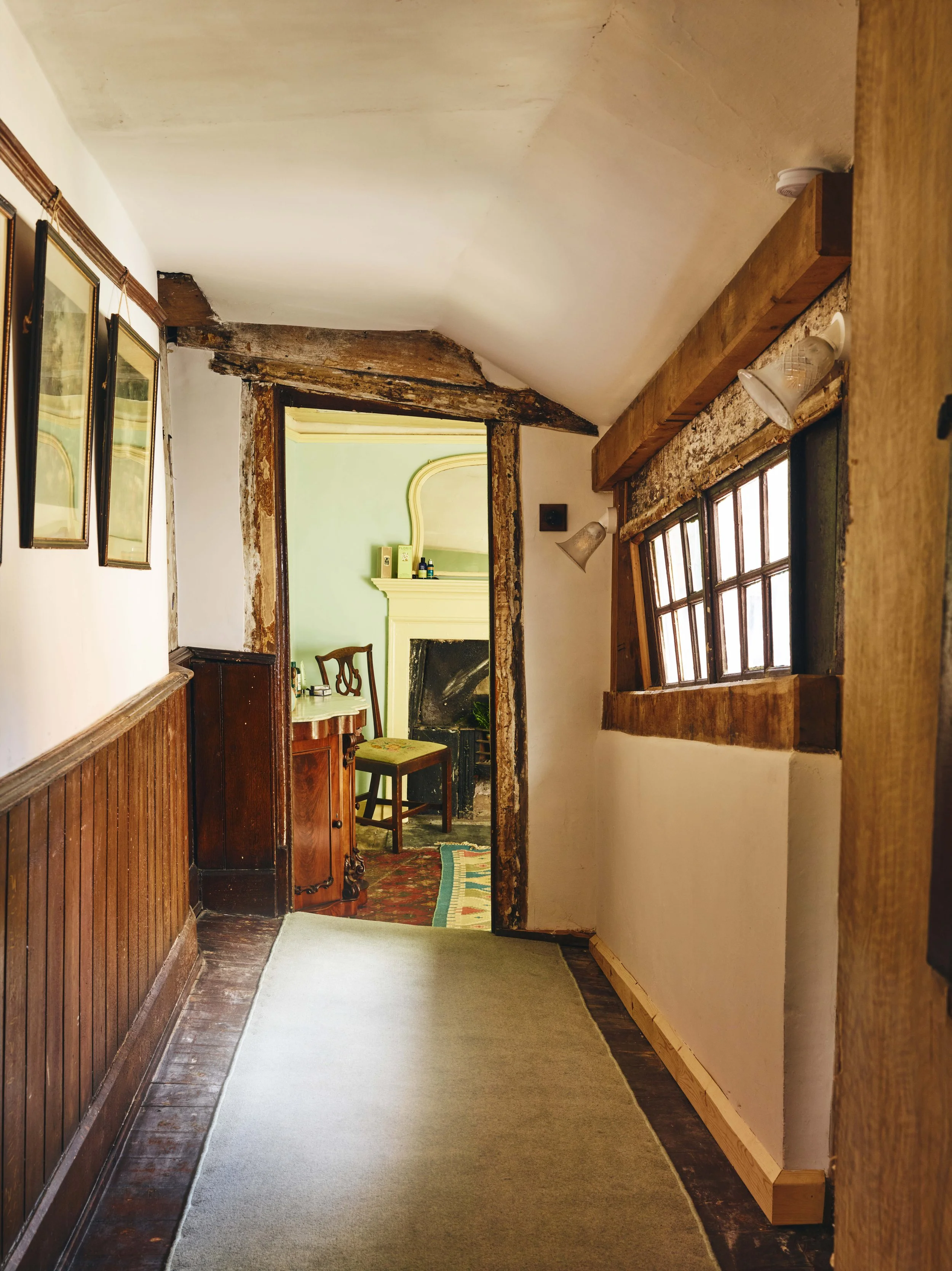 A hallway in a 16th century house in Cornwall with wooden accents leading to a green room with a fireplace and vintage furniture.