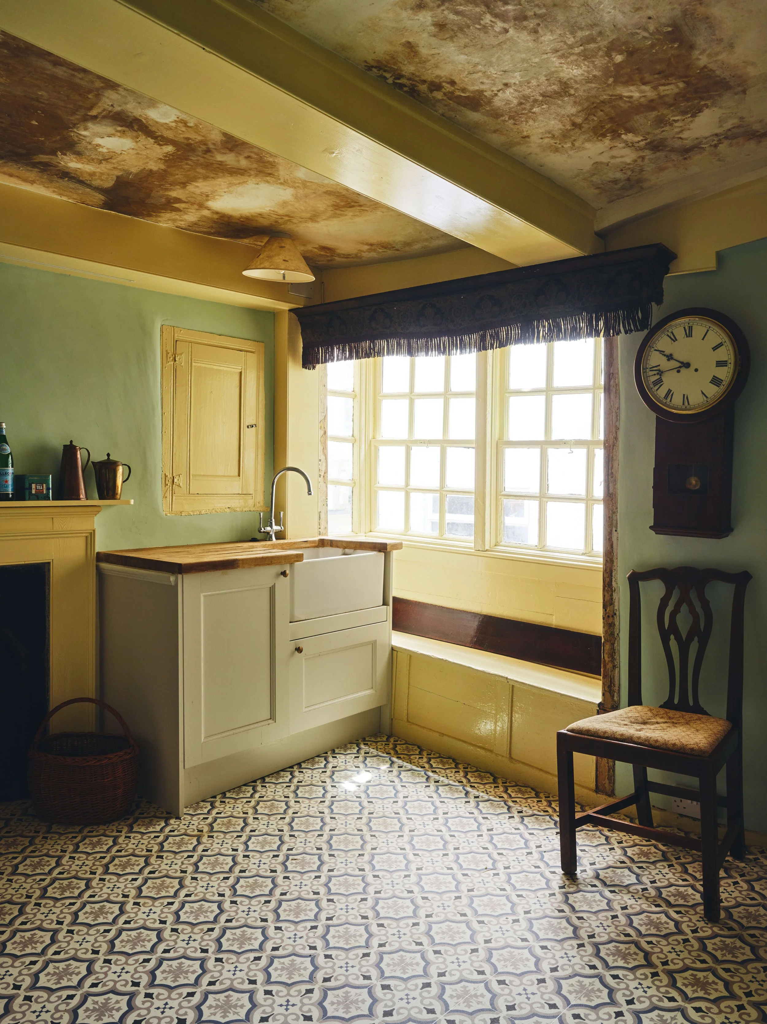 Historic kitchen in a 16th century house in Cornwall with patterned tile floor, green walls, large sunlight window, white sink cabinet, wall clock, wooden chair, and rustic ceiling.