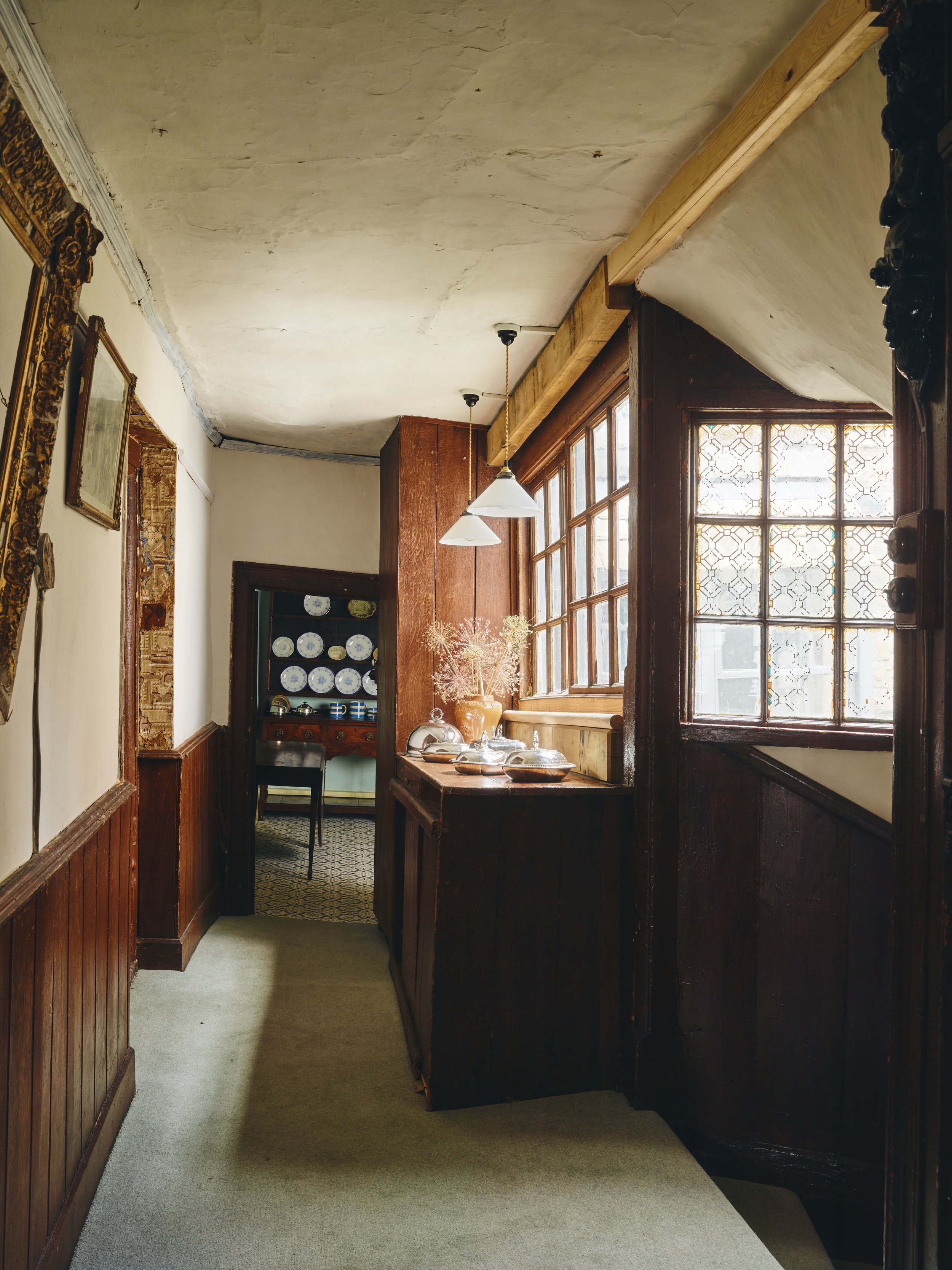 Narrow hallway with wooden paneling and a large window on the right, leading to a dining room with a table and chairs. Decor includes plates on a wall display, vases, and framed pictures.