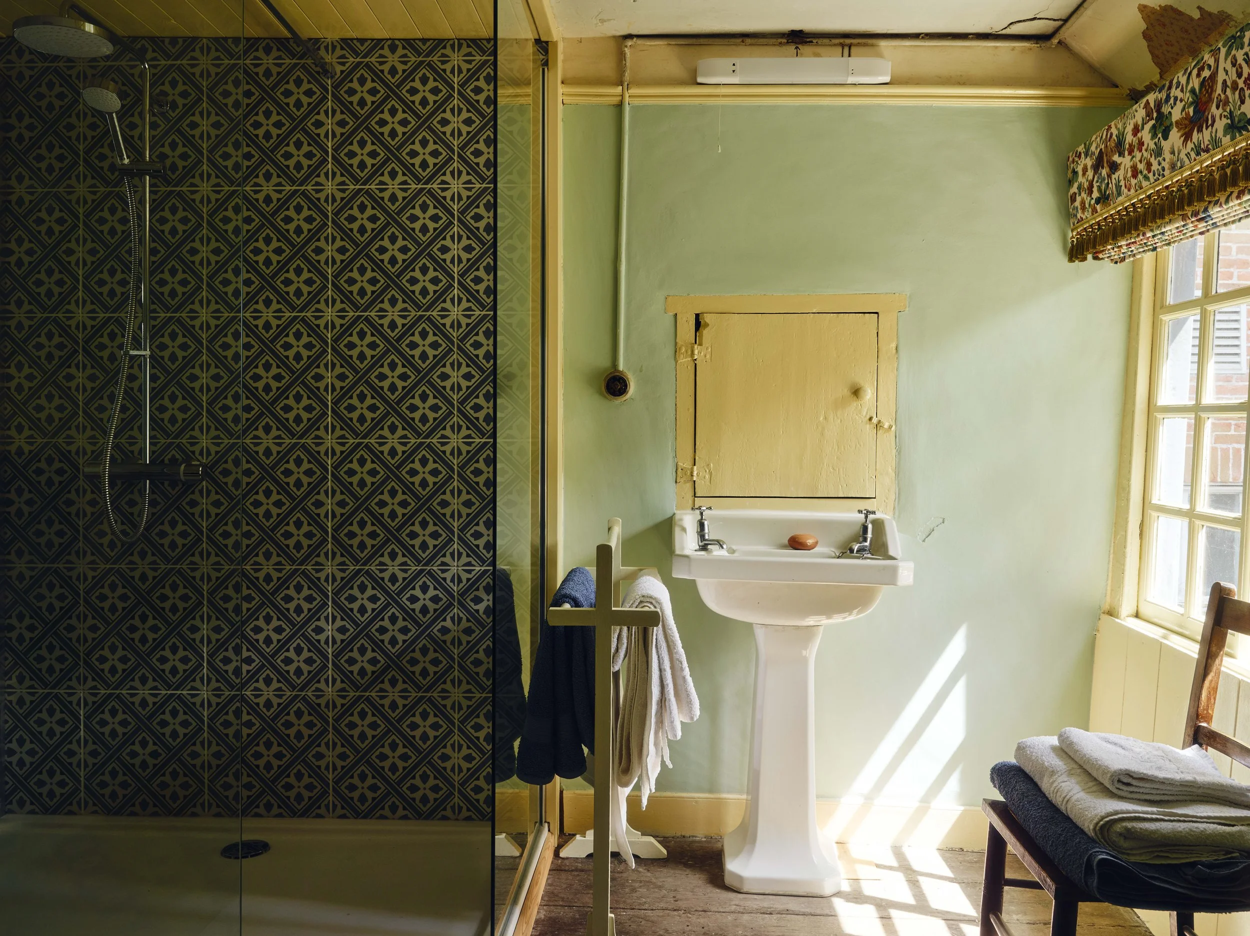 A bathroom with a vintage sink, a small wooden bench with folded towels, a window with floral curtains, and a separate shower area with dark patterned tiles.