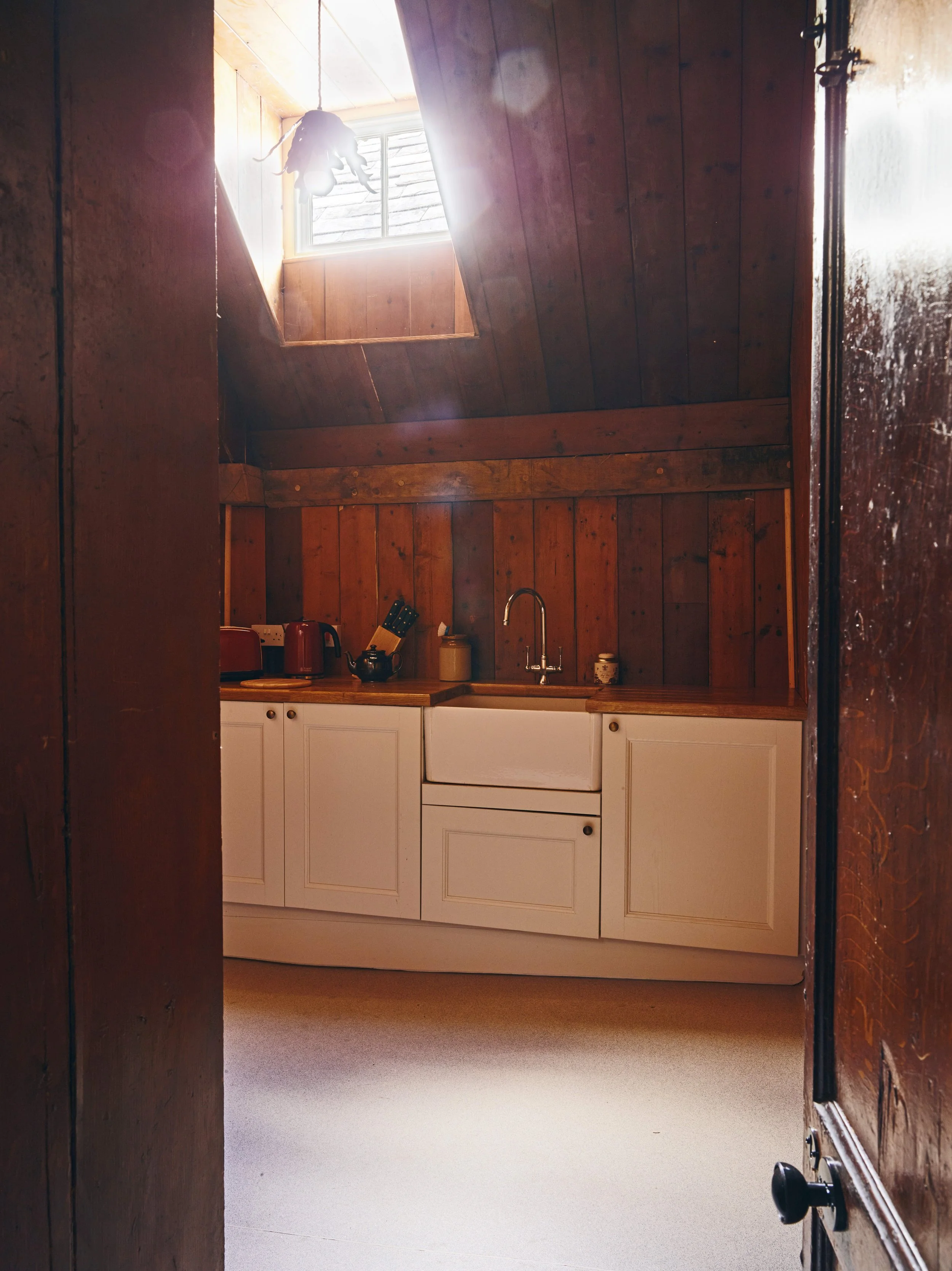A small kitchen with white cabinets, wooden countertops, and a wooden-paneled wall. There is a window with sunlight coming through and a sloped ceiling with a skylight. A hanging light fixture is visible in the upper part of the image.