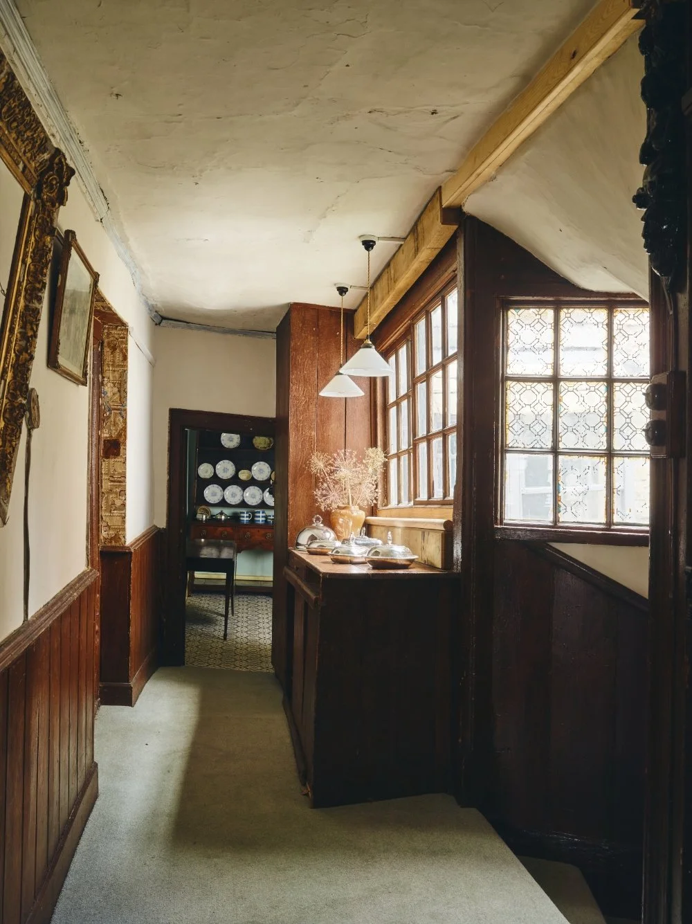 A hallway with wood paneling and large windows with decorative glass, leading to a dining room with a china cabinet filled with plates.