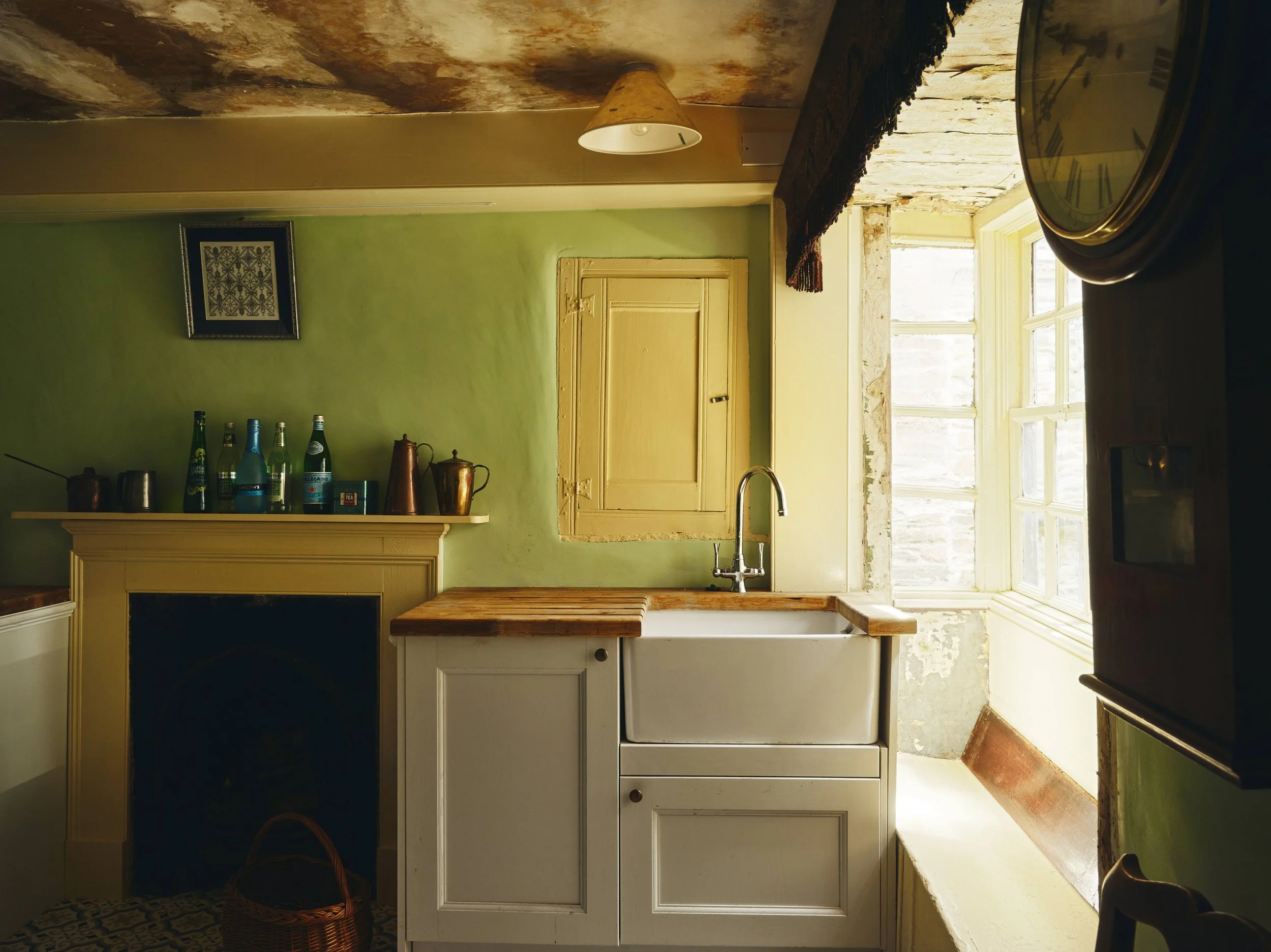 Historic kitchen with yellow cabinets, a farmhouse sink, green walls, a window with peeling paint, bottles and teapots on the mantle, a wall clock, and a basket on the floor.