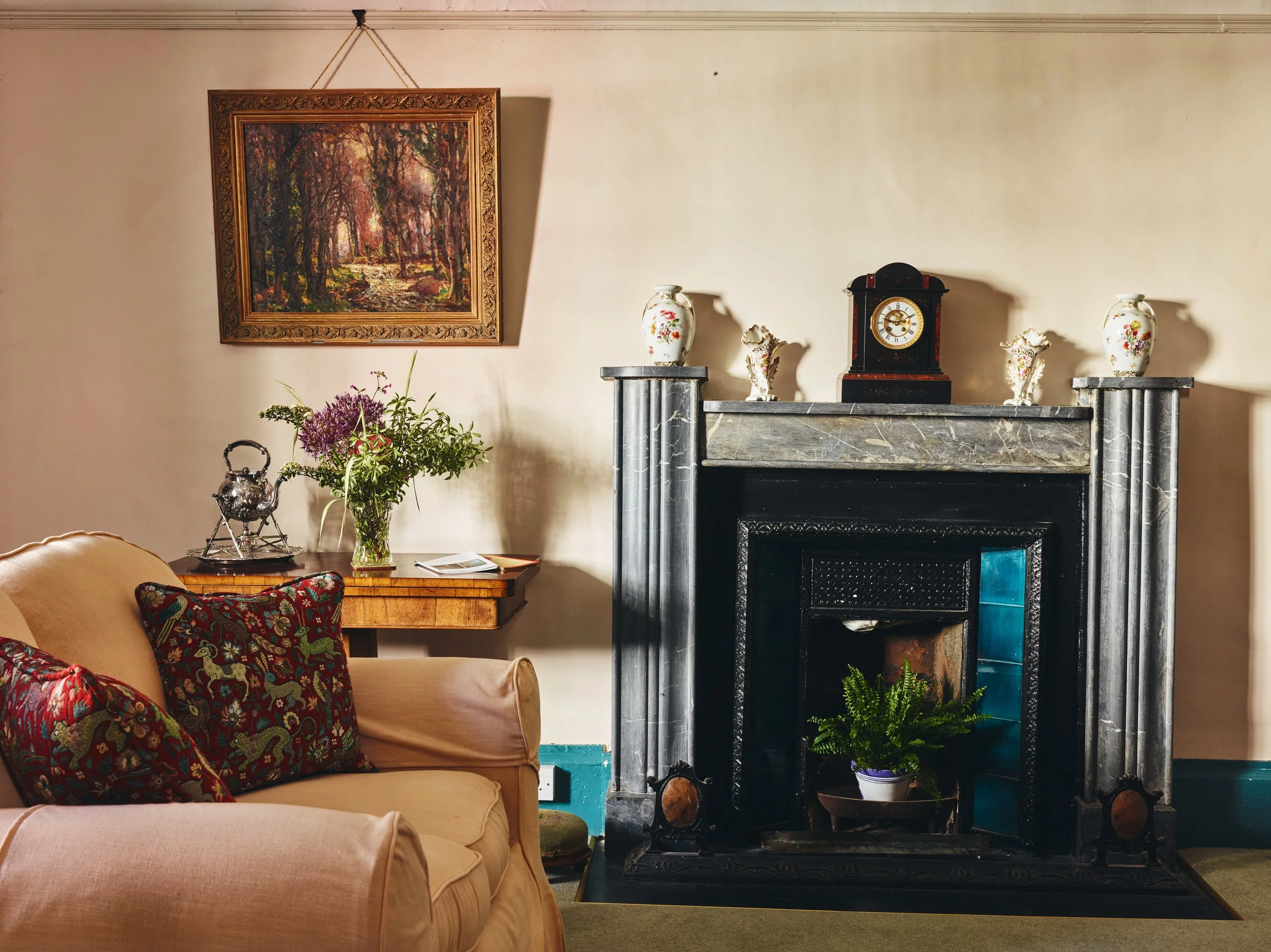 A living room with a fireplace featuring a marble mantel, decorative vases and a clock on top. To the left, a beige armchair with two patterned pillows, and a small wooden table with a vintage teapot, a vase of purple flowers, and a book. A framed la