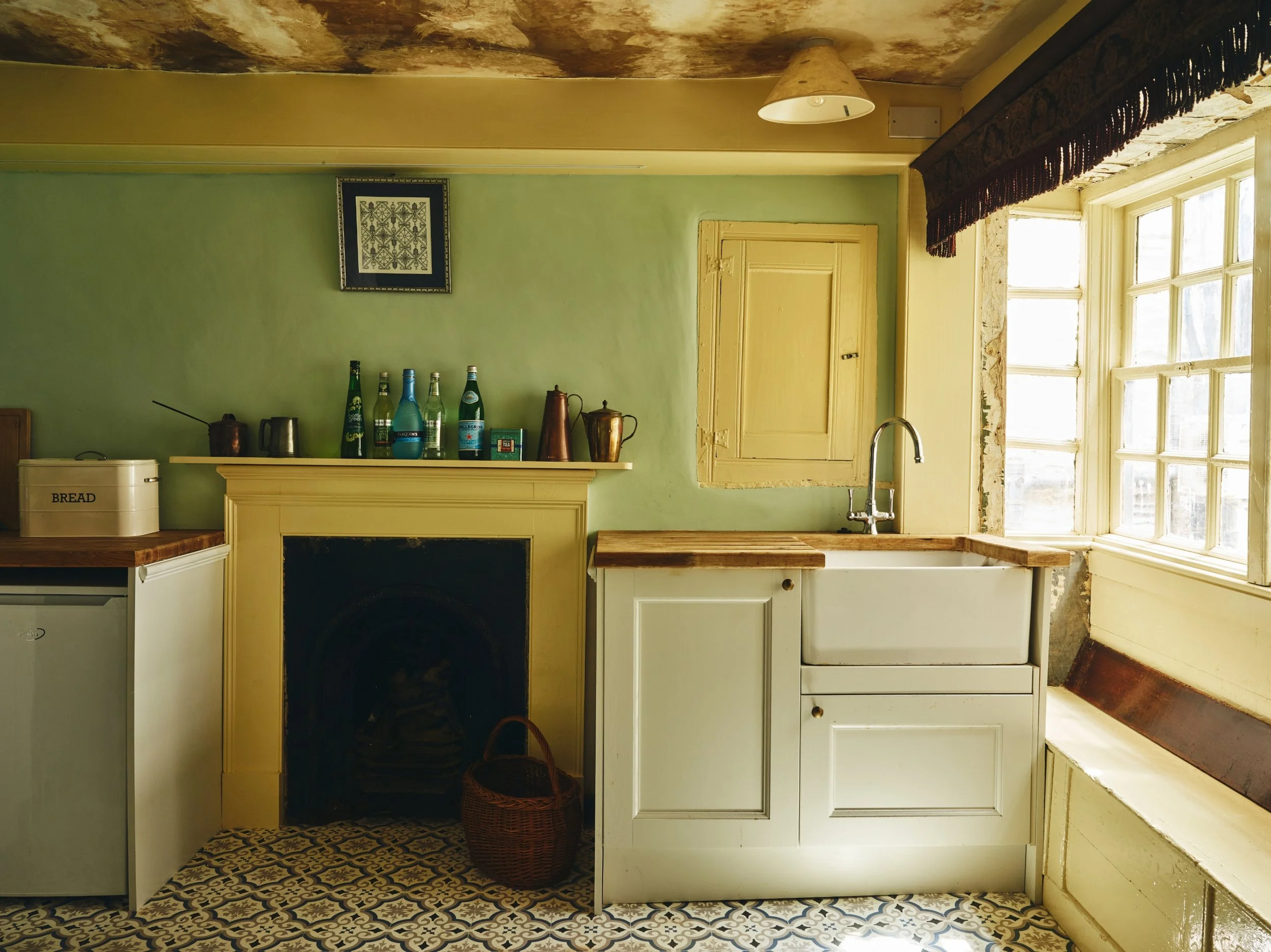 A  kitchen with a green wall, a yellow wooden fireplace mantel with bottles on top, a white sink with a wooden countertop, a small closed yellow cabinet above the sink, a window with mullions, a white cabinet with a basket in front, and patterned floor tiles.