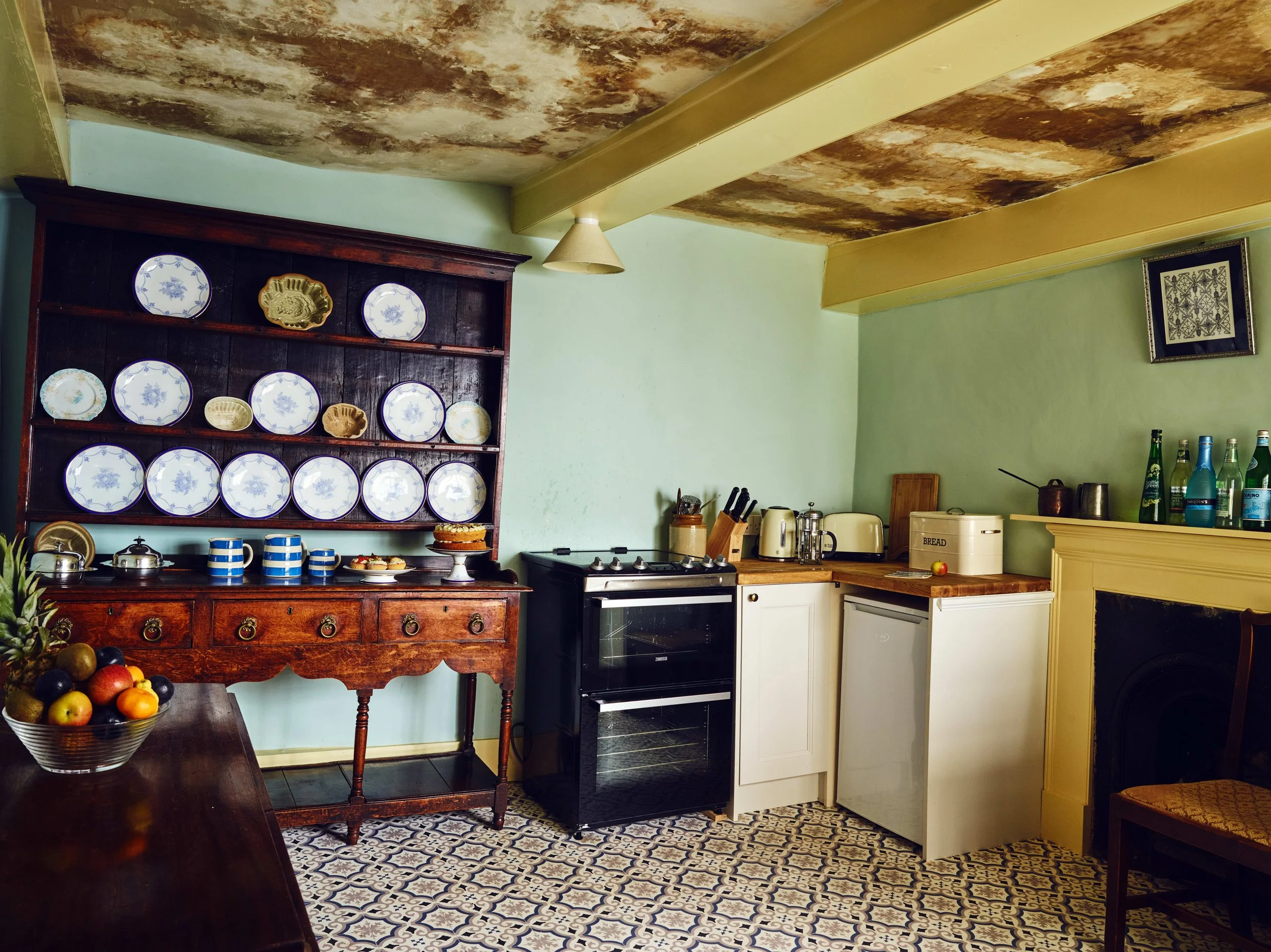 Kitchen with a dark wooden hutch displaying decorative plates and dishes, a patterned tiled floor, light green walls, and a mixture of modern and vintage appliances including a stove, mini fridge, and toaster. A bowl of fruit is on the table.