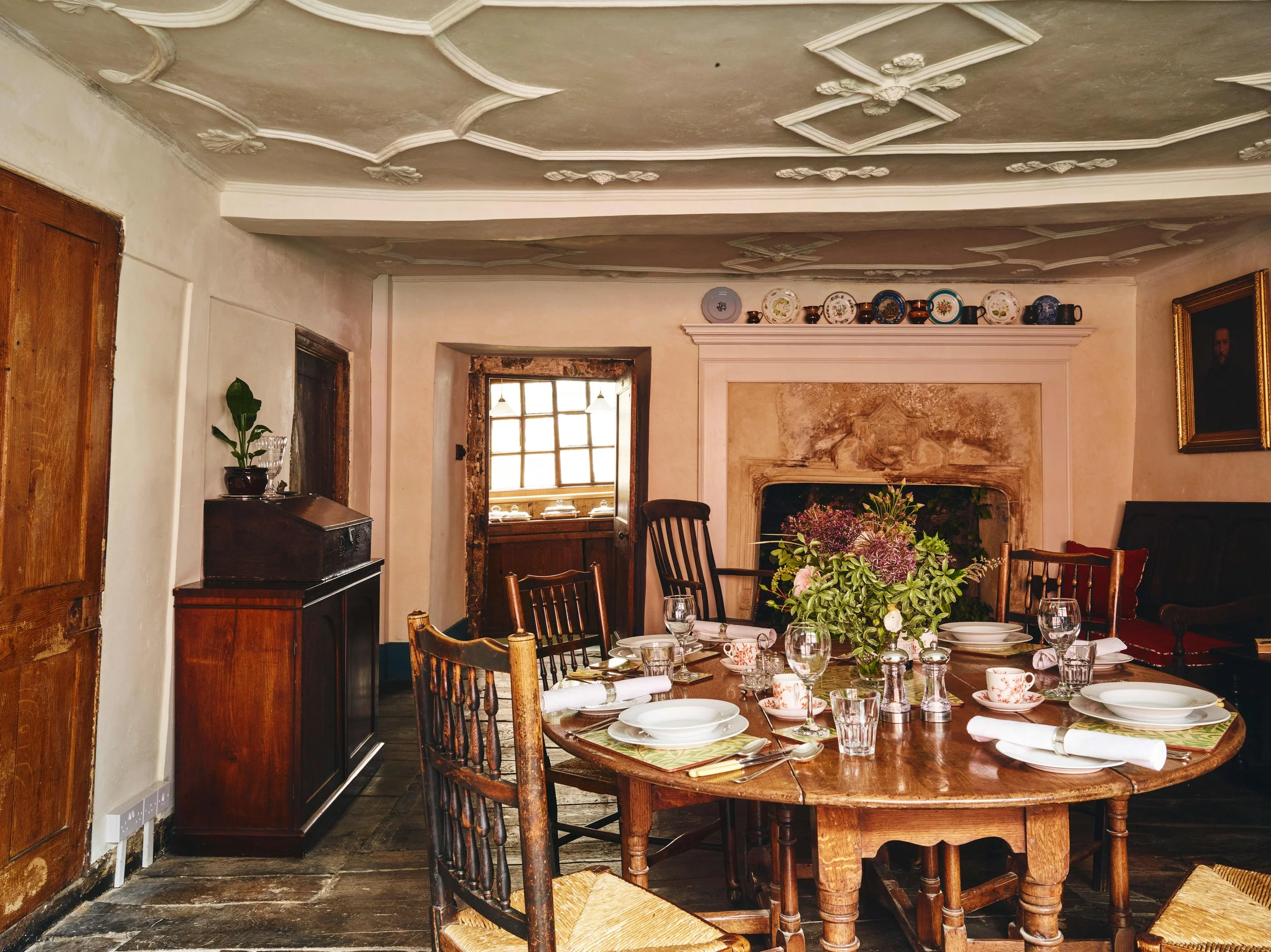 A  dining room with a round wooden table set for a meal, featuring white plates, cups, glasses, and silverware, with a large floral centerpiece. The room has a vintage style with wooden chairs, a fireplace, a cabinet, a window, and decorative plates 