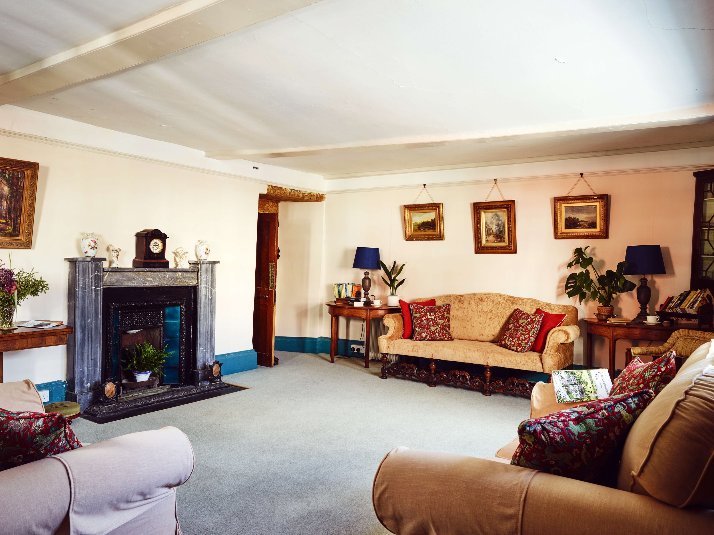 A living room with a beige sofa, armchairs, framed paintings, a fireplace, and side tables with lamps and decorations.