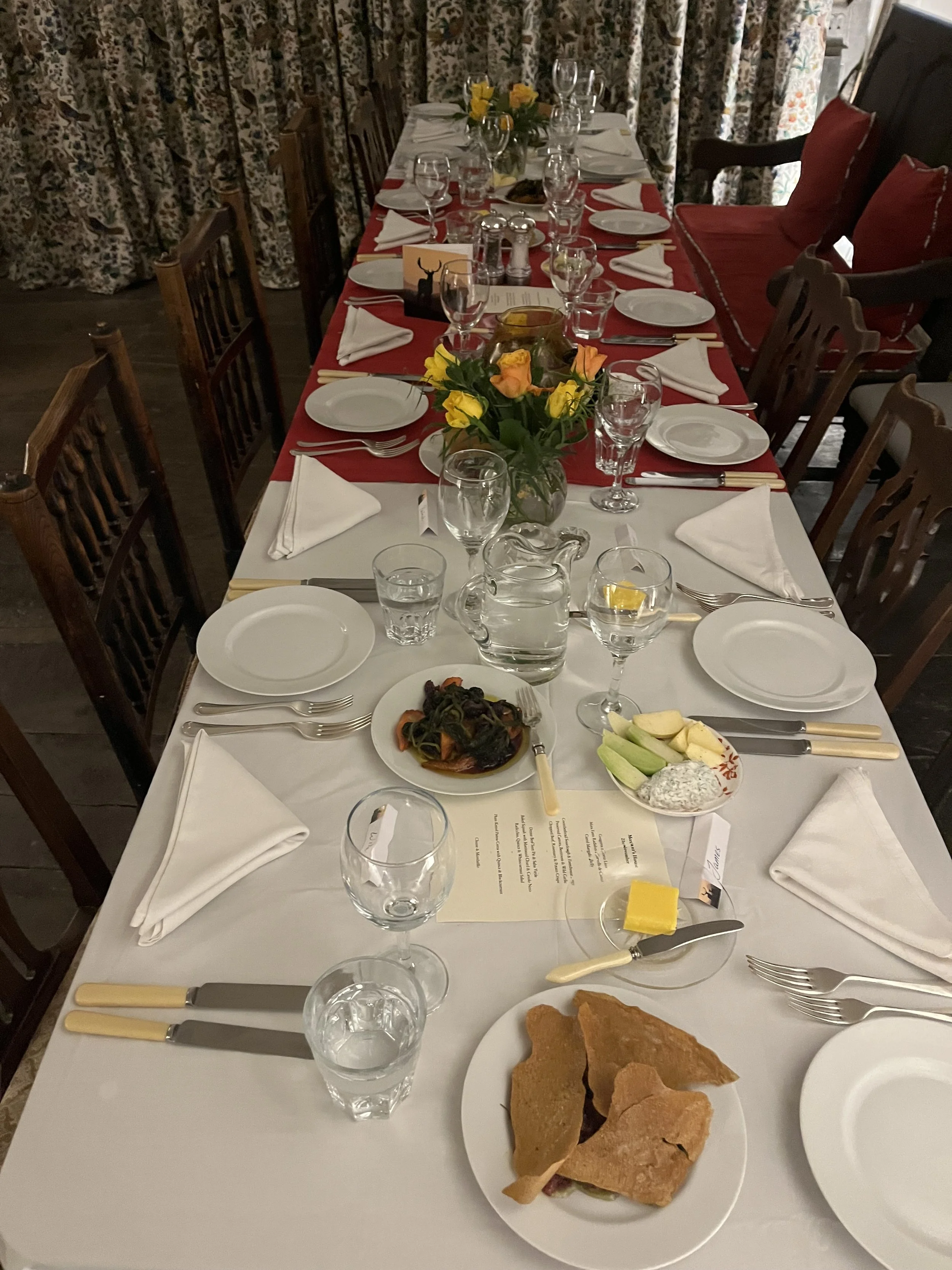 A long dining table set for a meal with white tablecloths, floral centerpieces, place settings with white napkins, glassware, and silverware. There are some plates of food. The background shows curtains with a floral pattern and wooden chairs around the table.