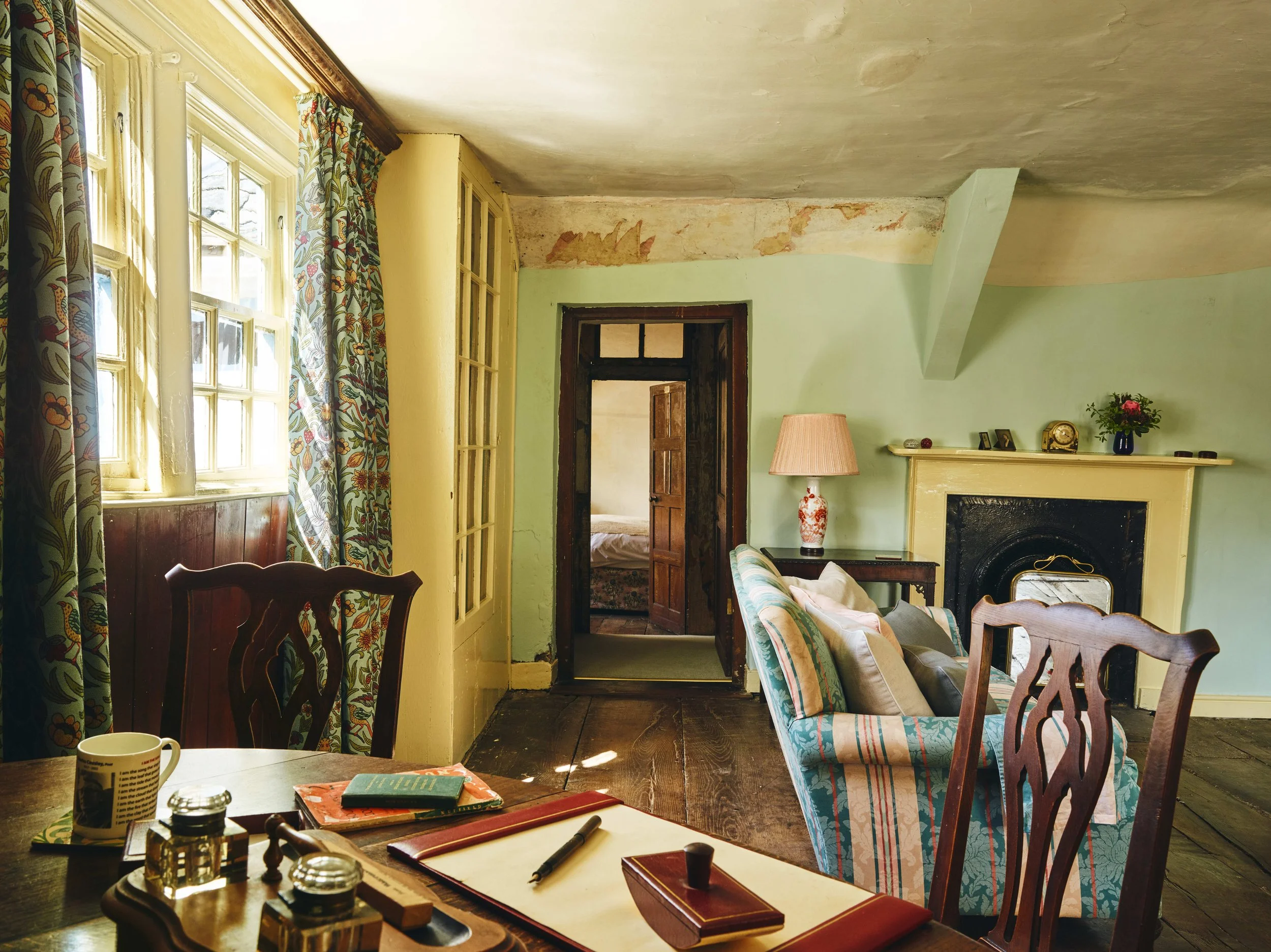 Historic living room in a16th century house in Cornwall with a wooden dining table, decorative chairs, a sofa with cushions, a fireplace, and a lamp. Sunlight comes through patterned curtains on windows.