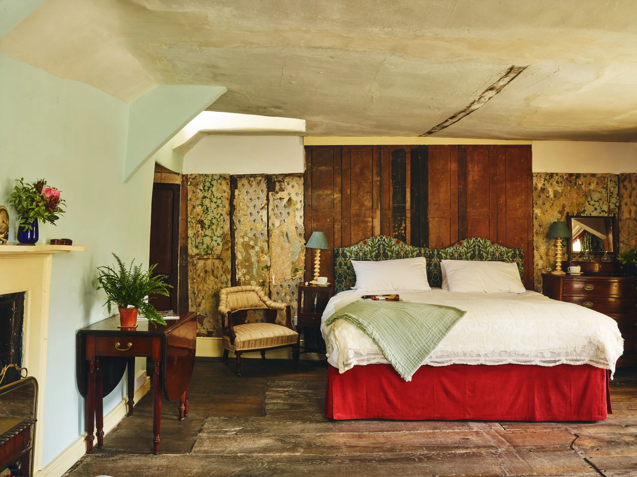 A bedroom with historic decor, a large bed with floral headboard, white bedding, green blanket, and red bed skirt, wooden furniture, lamps, chairs, plants, and a fireplace.