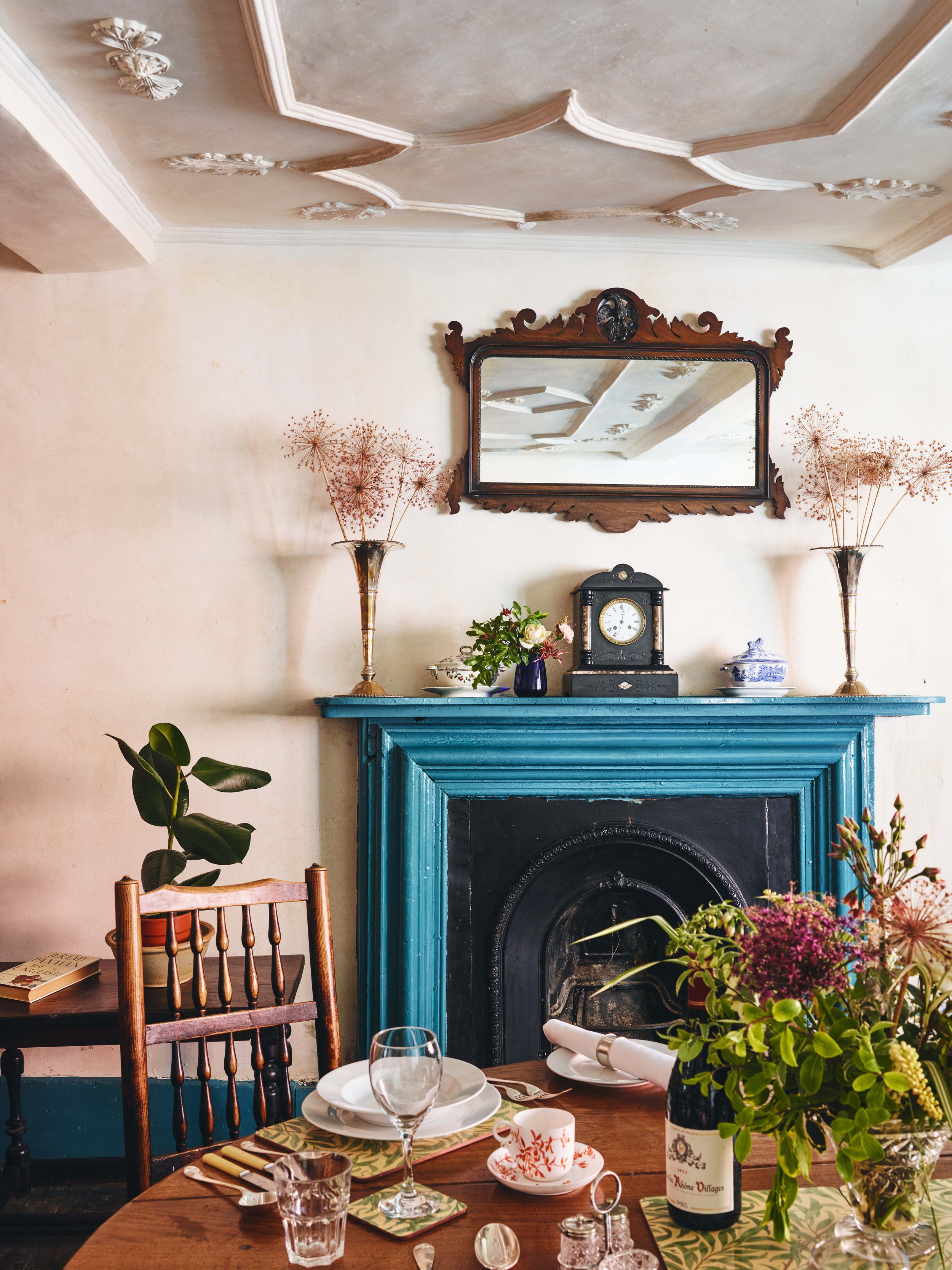 A historic dining room with a teal fireplace mantel, decorated with flowers, vintage clock, and a mirror, with a table set with dishes and cutlery, and plants and vases on the mantel and nearby.