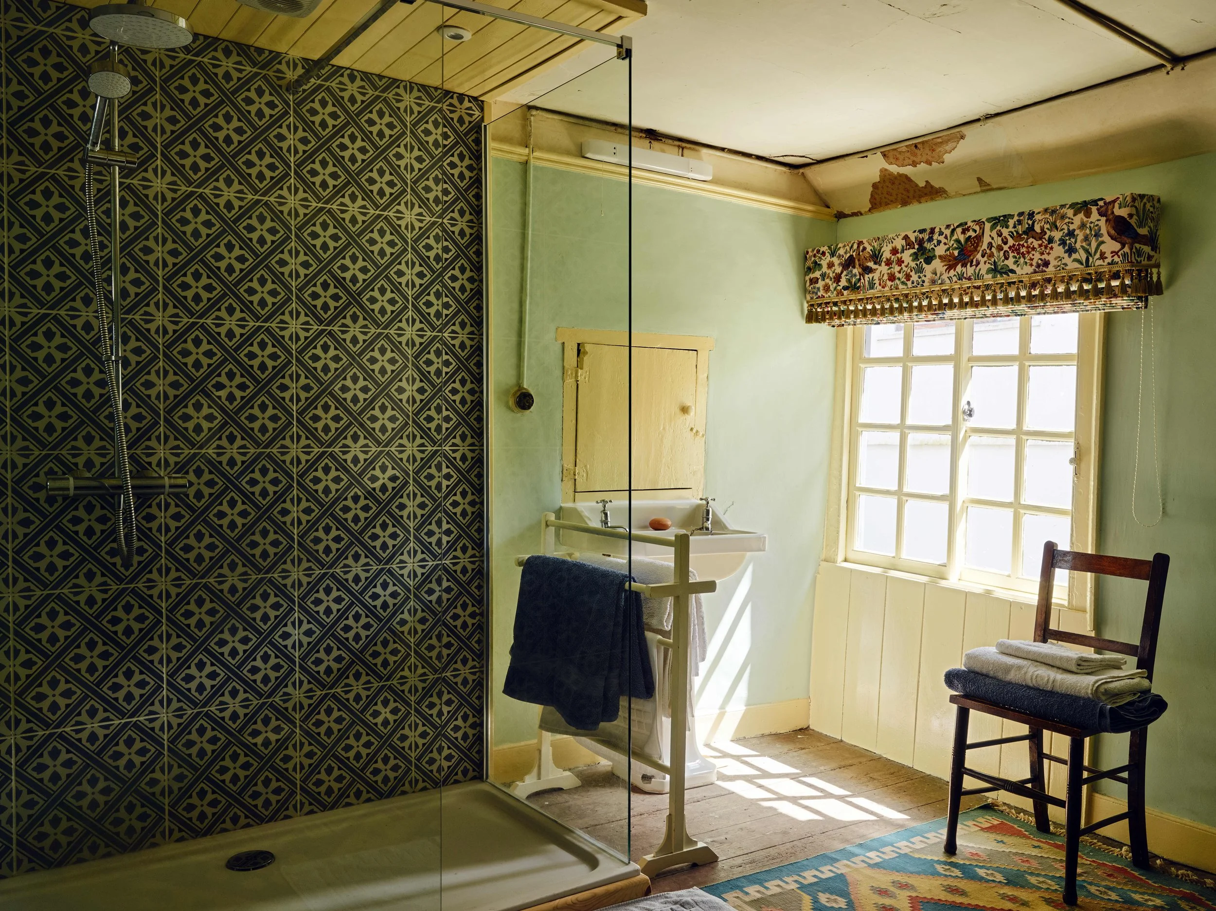 Bright bathroom with a tiled shower, vintage sink on a stand, wooden chair with folded towels, window with floral valance, green walls, peeling ceiling paint, patterned rug.