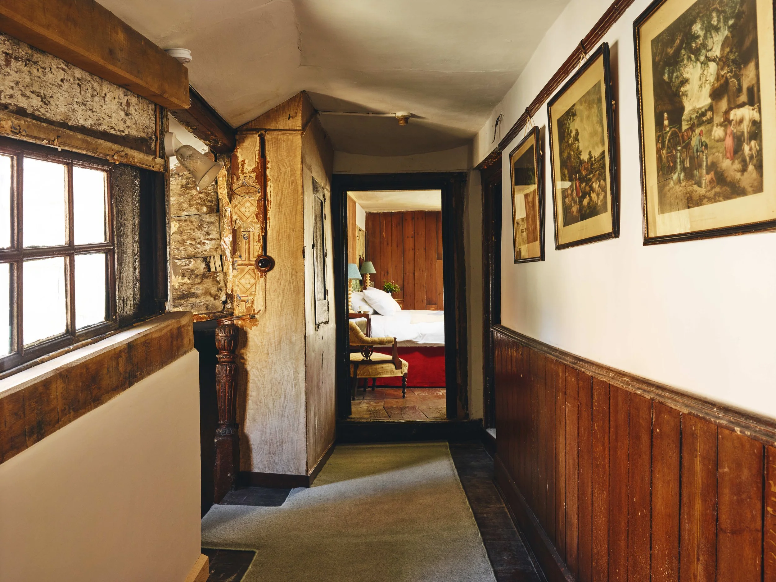 A narrow hallway with wood-paneled walls and a beige carpet leading to a bedroom with a bed, nightstands, and lamps.