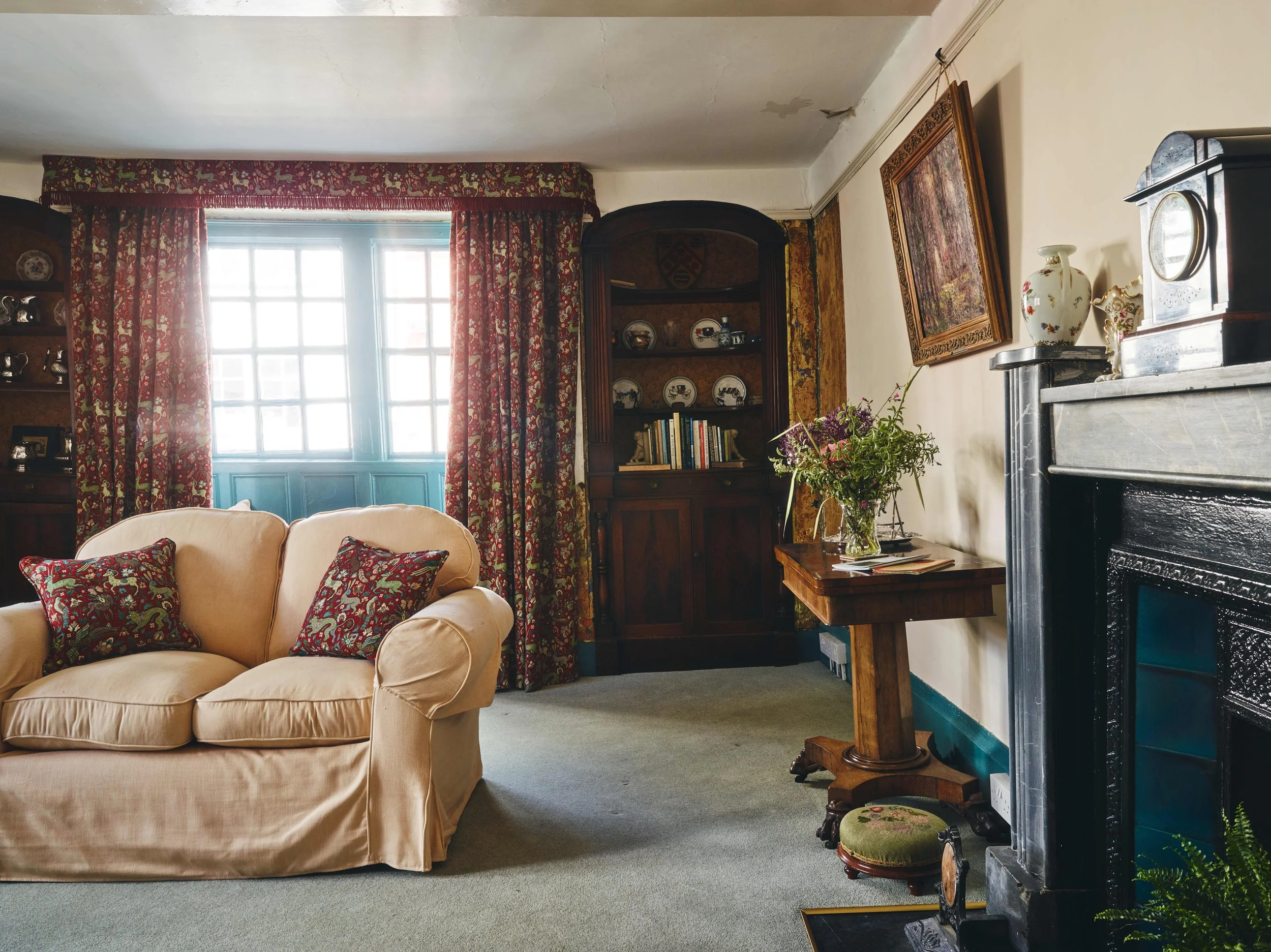 Living room with a beige sofa, red patterned curtains, a wooden cabinet with books and dishes, a small wooden table with a vase of flowers, a black fireplace, and framed artwork on the wall.
