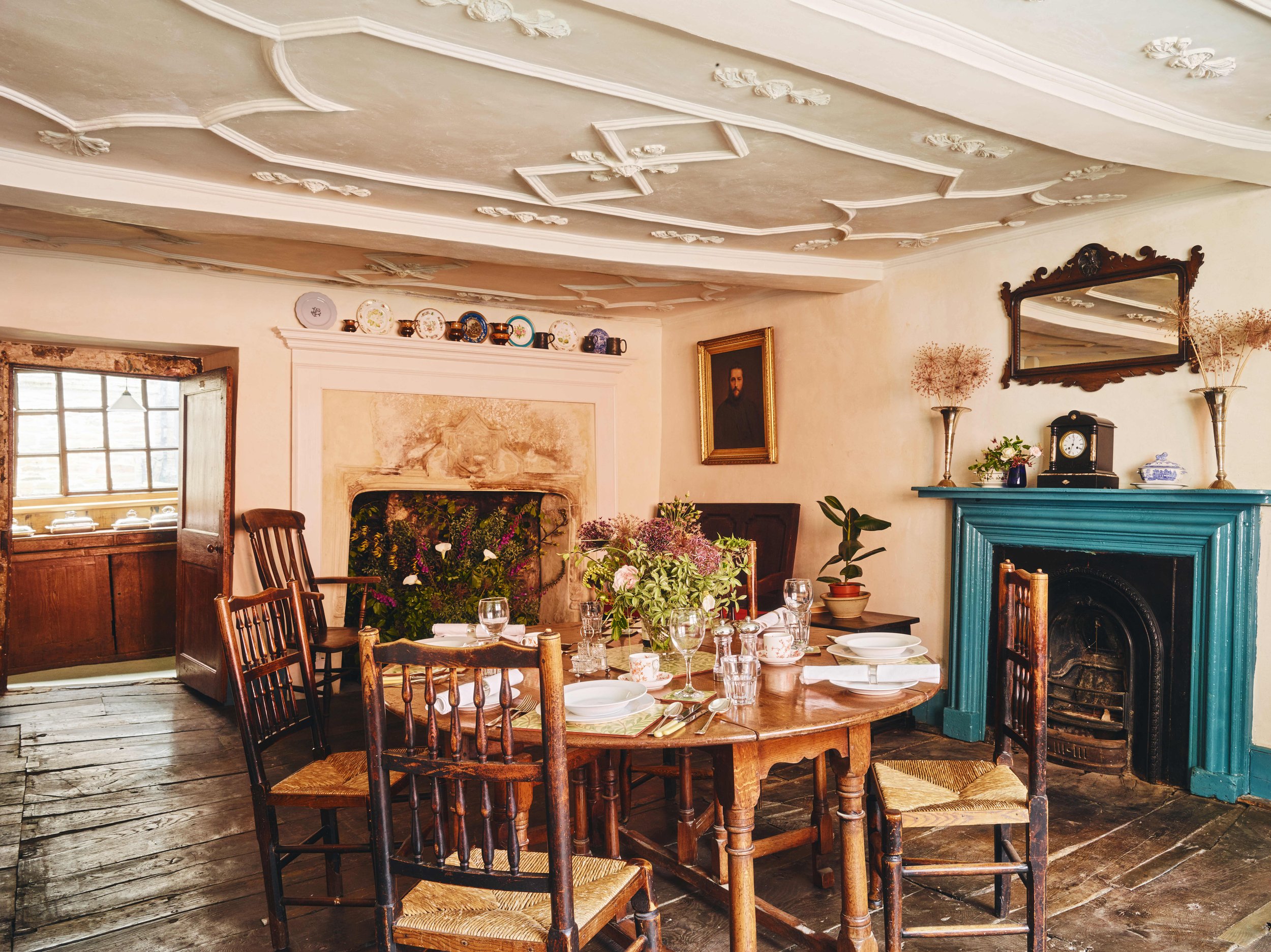 A dining room with a round wooden table set for a meal, adorned with a floral centerpiece, surrounded by wooden chairs, a fireplace with a turquoise mantel, decorative plates on the shelf above, a portrait on the wall, and a mirror reflecting part of