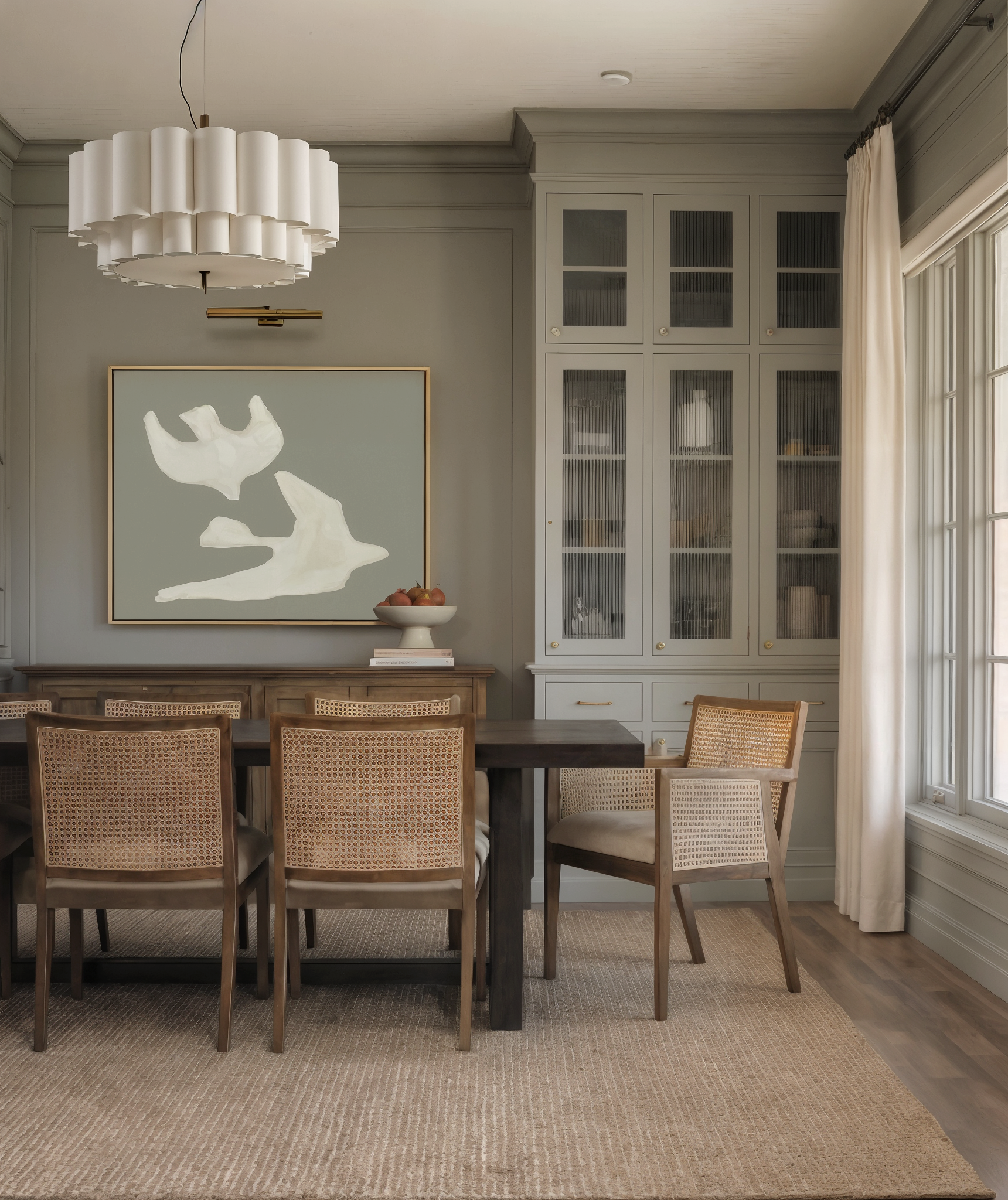Transitional dining space featuring muted green built-in cabinetry, cane dining chairs, and a tiered fabric pendant light.