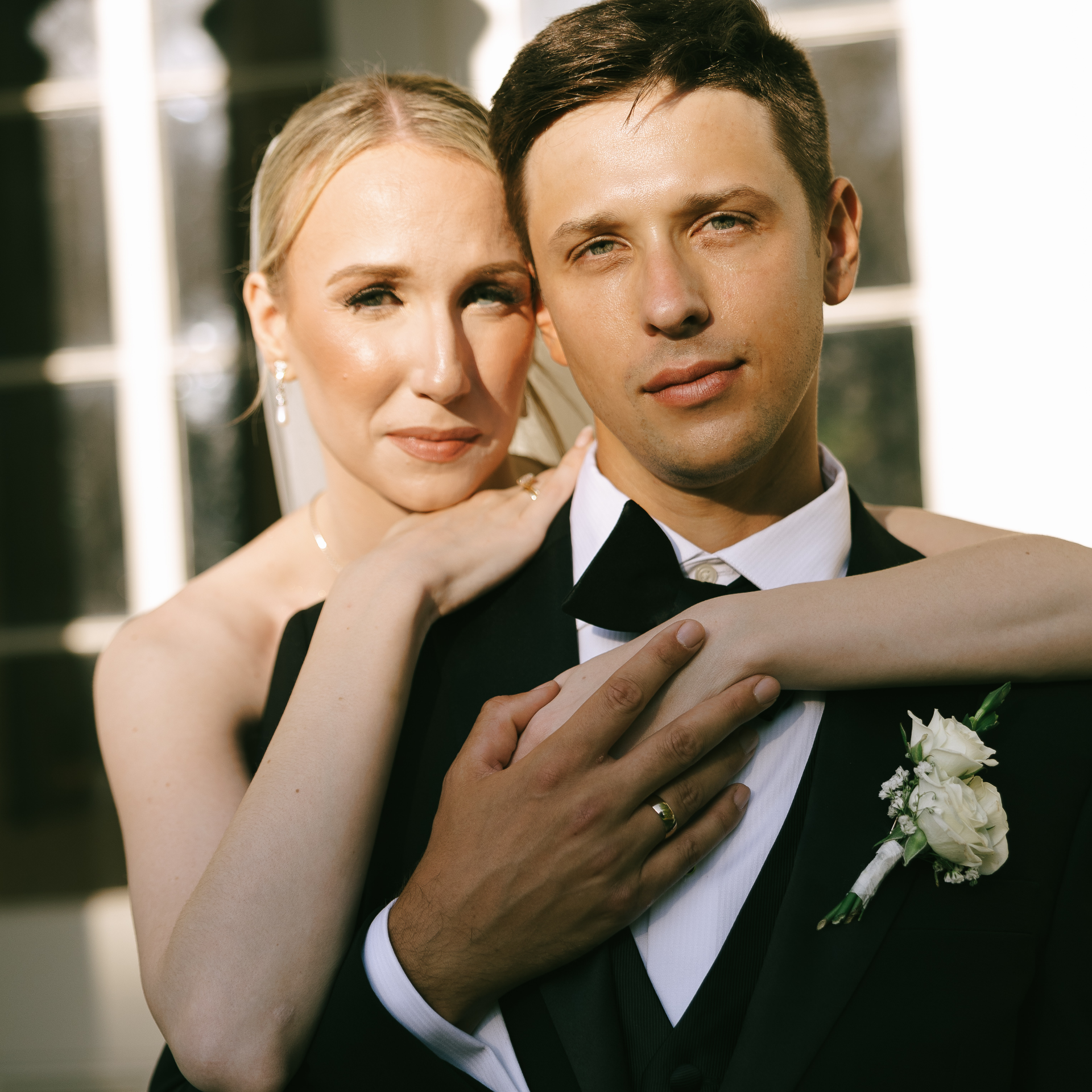 A bride and groom on their wedding day, with the bride embracing the groom from behind, outdoors with white framing and greenery in the background.