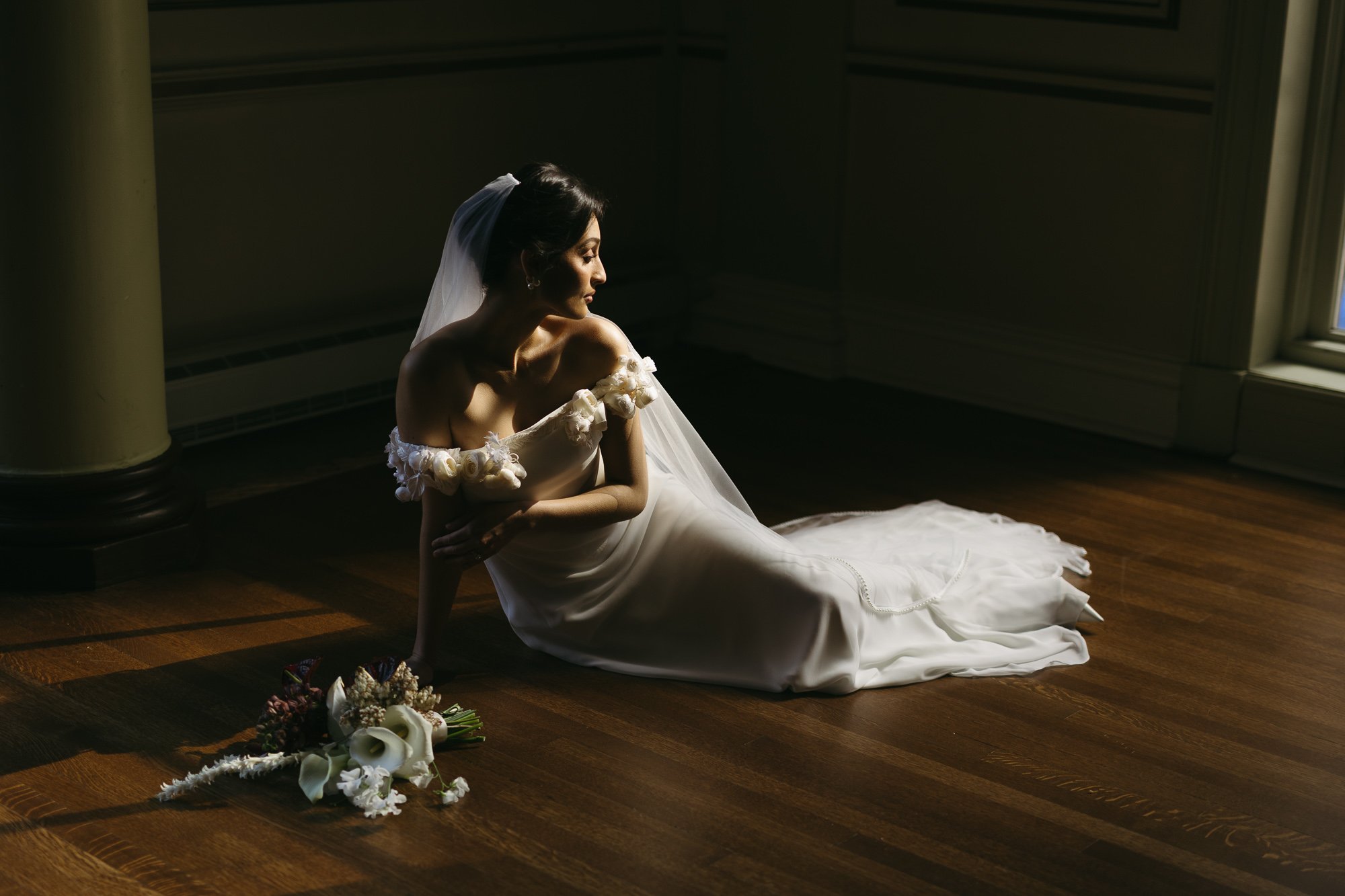 A woman in a wedding dress sitting on a wooden floor near a window with sunlight coming in, with a bouquet of flowers beside her.