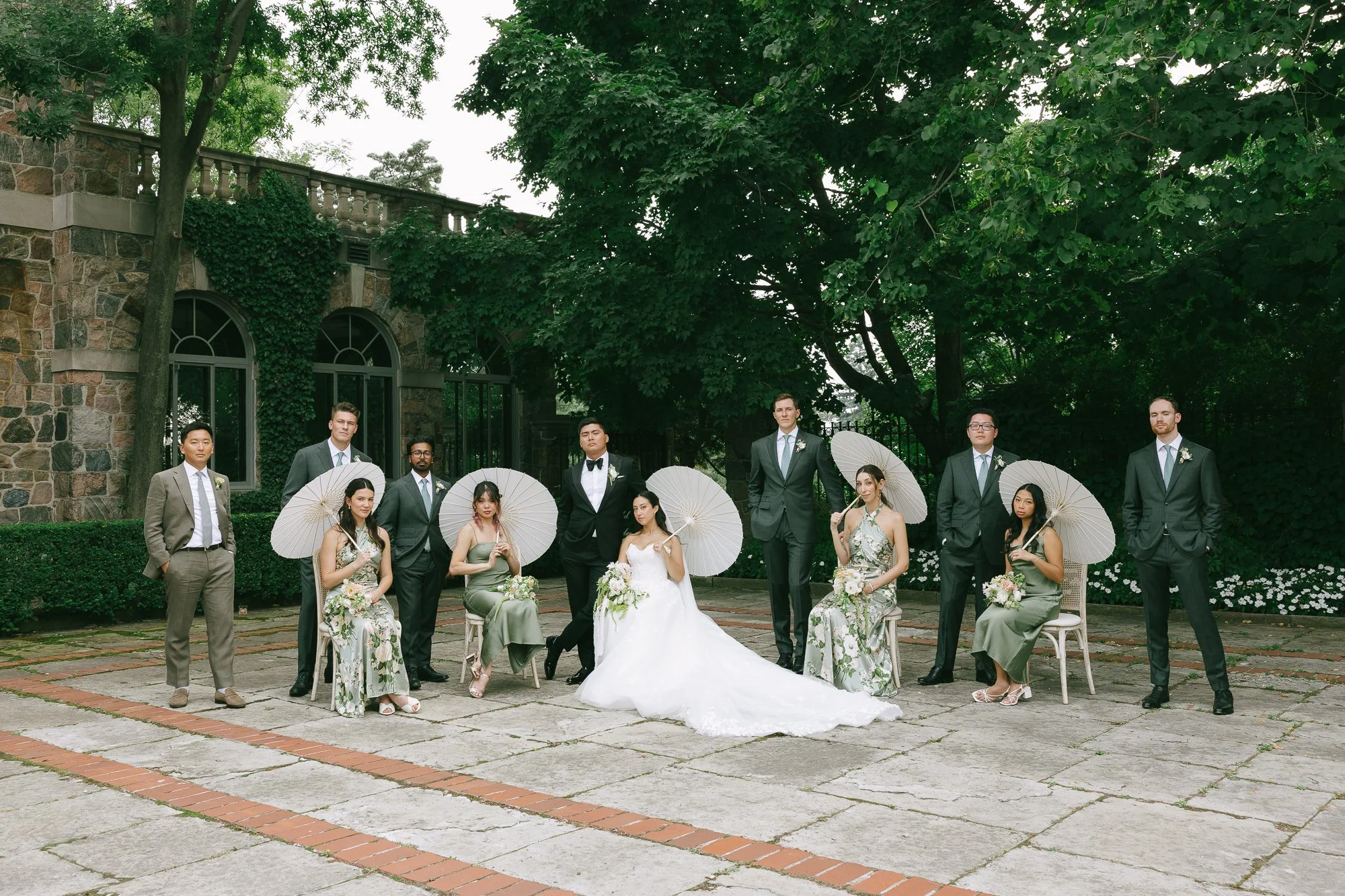 A wedding party poses outdoors on a brick and stone paved area, with a stone building and lush green trees in the background. The group includes the bride and groom, four bridesmaids, and five groomsmen, some holding white parasols.