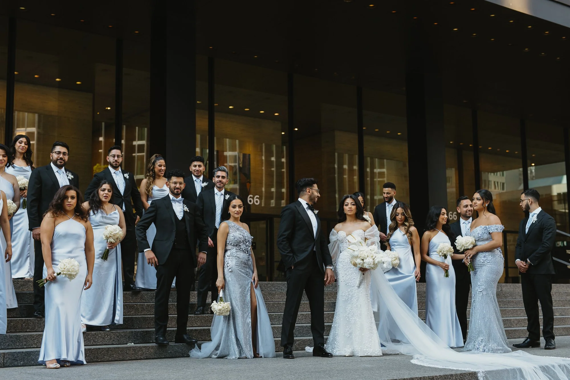 Group of wedding couples standing on steps in front of a modern building, dressed in formal wedding attire.