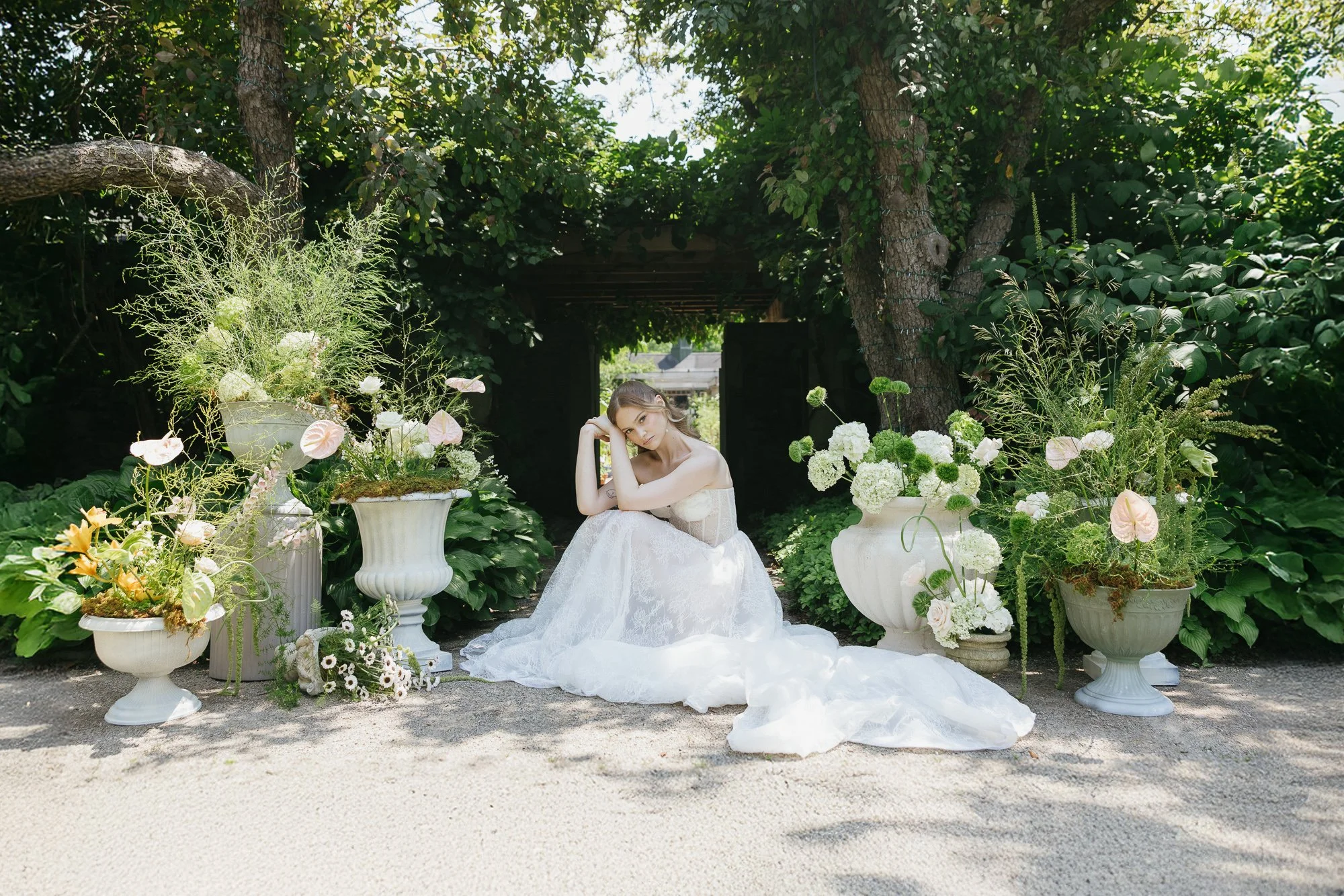 A woman in a white lace dress sitting on the ground amidst large white flower planters in a lush green garden.